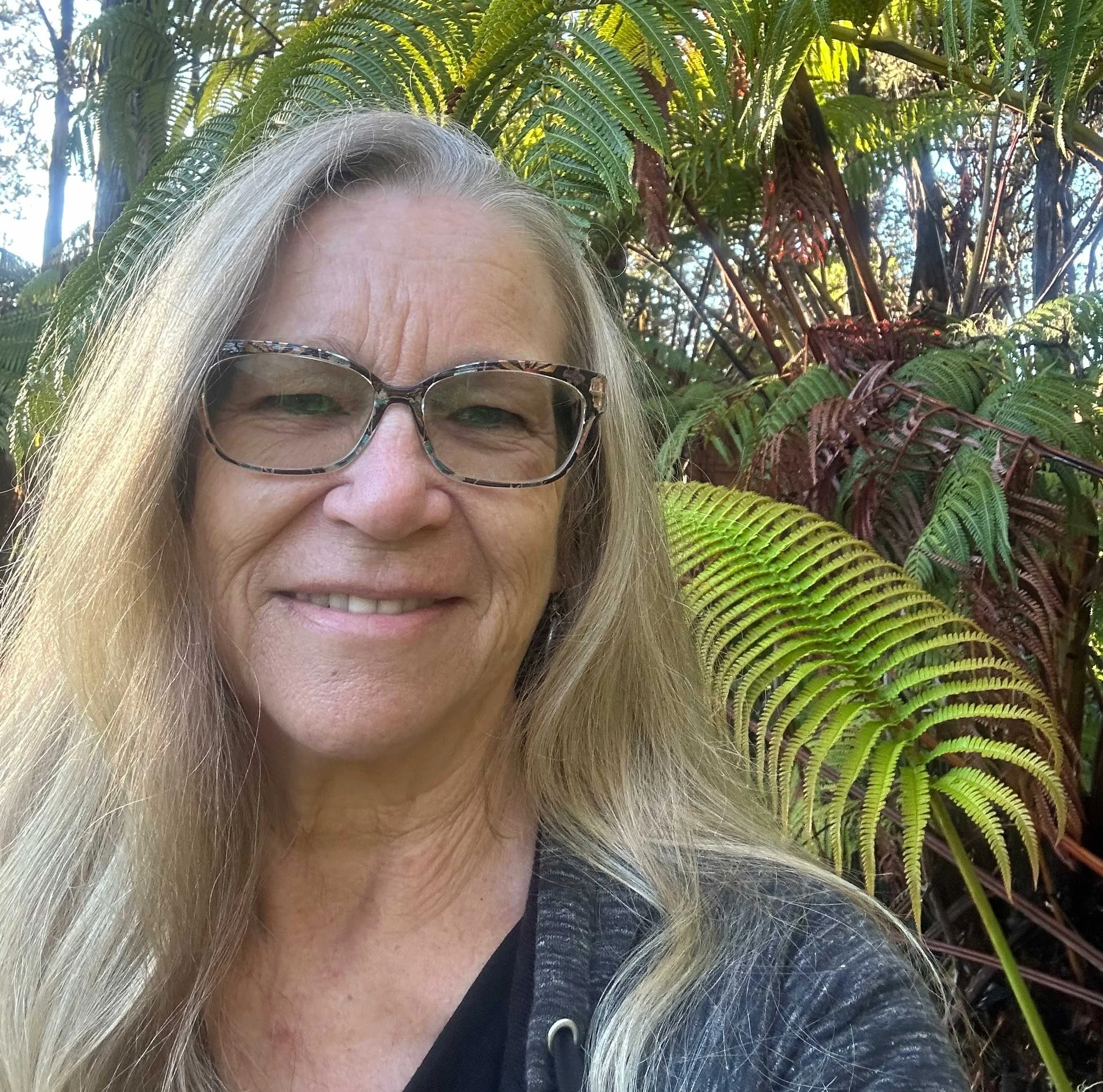 Partial view of a woman with long blonde hair, wearing sunglasses, standing outdoors with lush green ferns in the background.