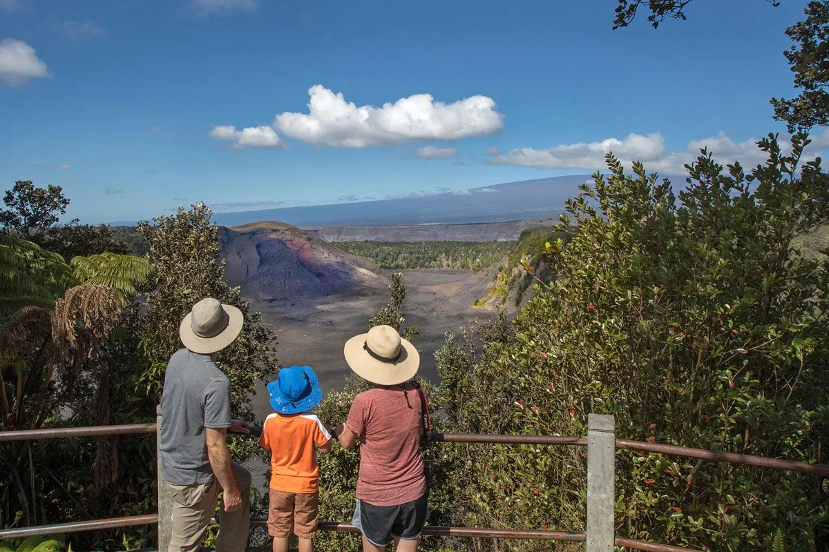 Three people, two adults and one child, standing on a viewing platform looking at volcanic landscape with clouds in the sky.