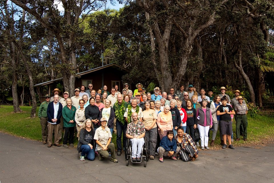 A large group of diverse people gathered outdoors, standing in front of a wooded area and a small building, posing for a group photo on a paved path.