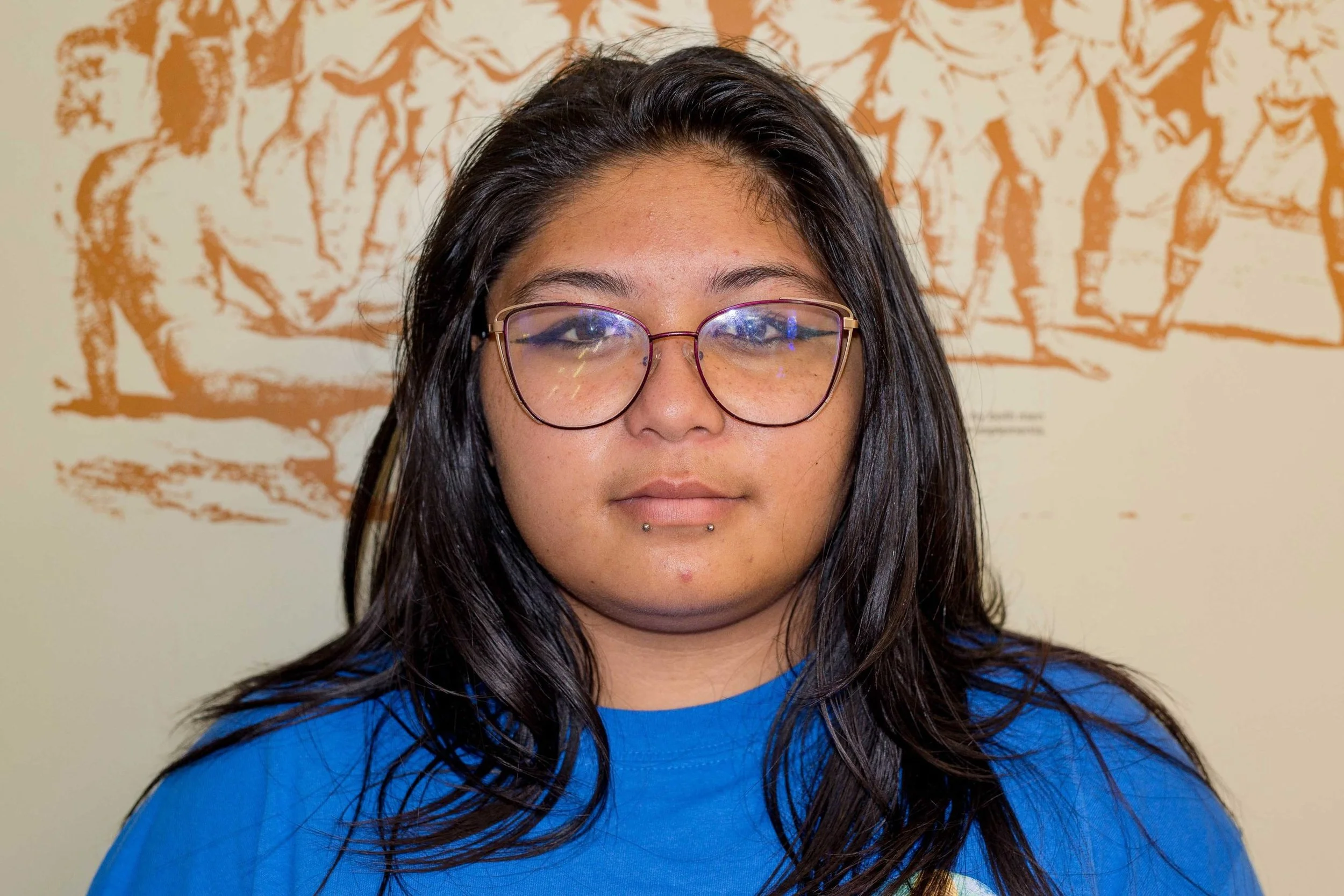 A young woman with long dark hair, wearing glasses and a blue shirt, standing in front of a wall with orange and white artwork.