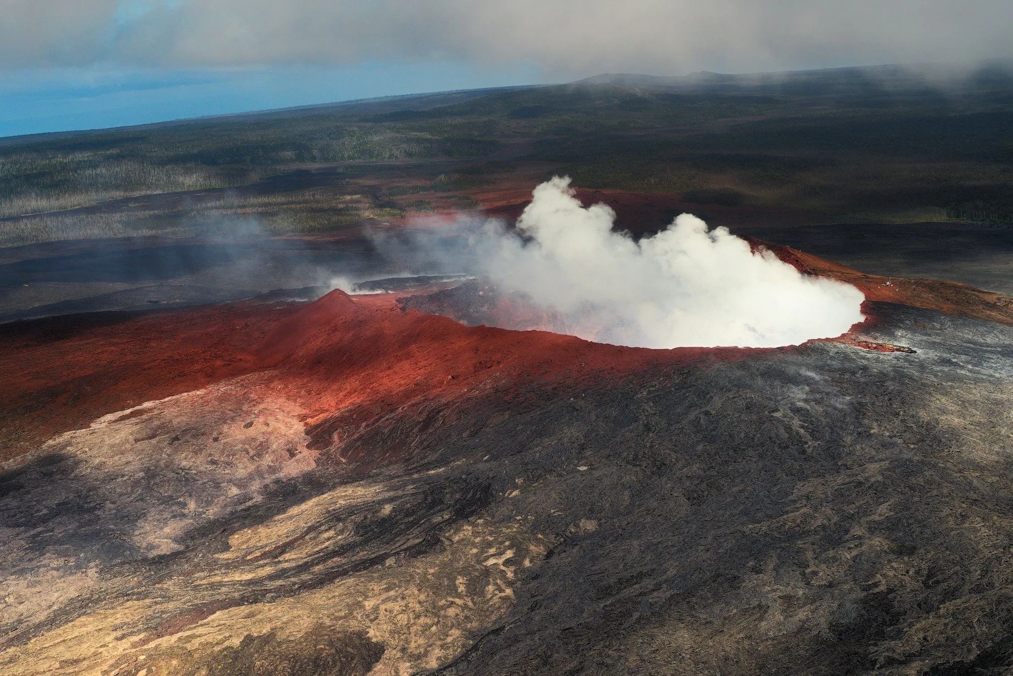 A volcanic landscape with red and black lava, steaming vents, and a misty background of forested mountains and a cloudy sky.