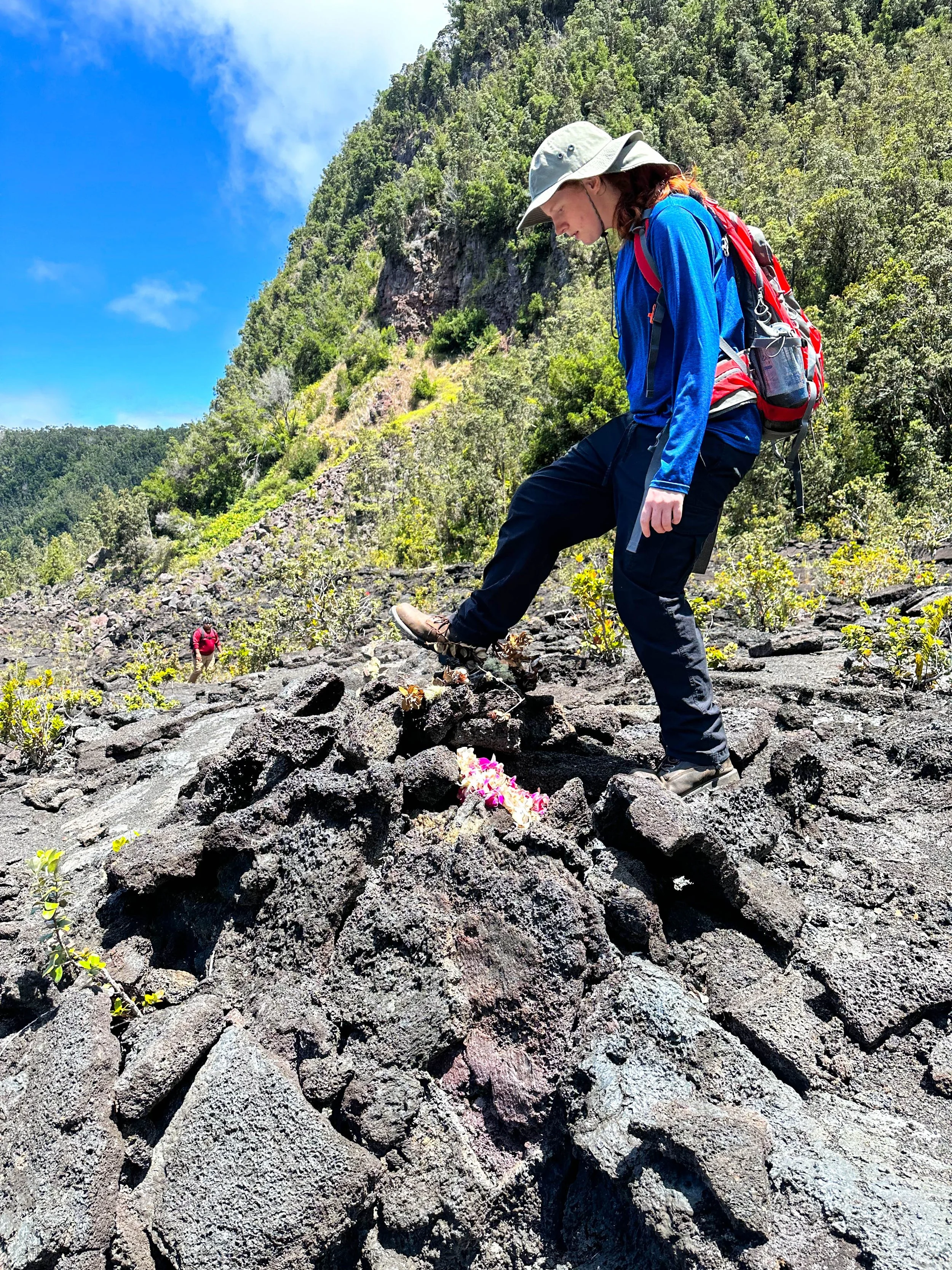 Hiker in blue jacket and gray hat placing a flower on volcanic rock with green forest and blue sky in background.