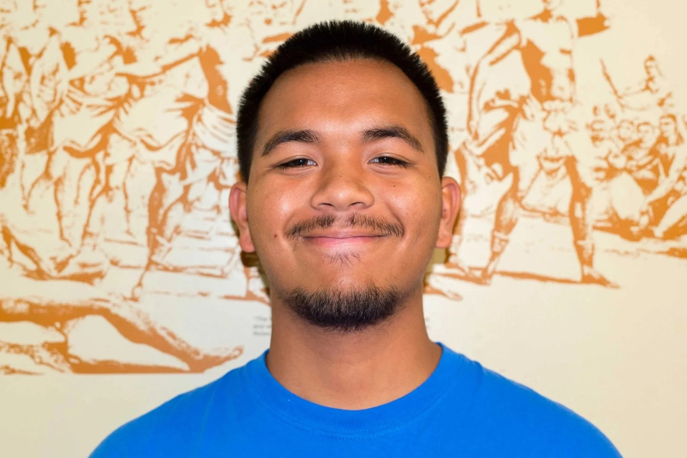 Close-up of a smiling young man with short black hair, a goatee, wearing a blue shirt, standing in front of an orange and white tree-patterned background.