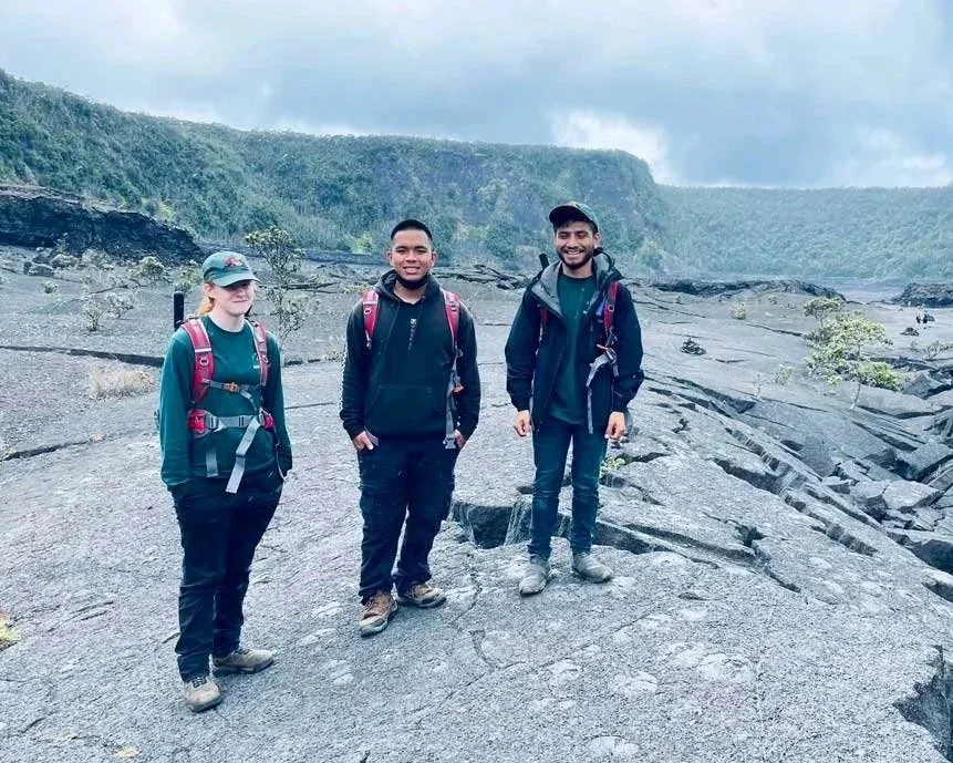 Three hikers standing on volcanic rock with green hills and cloudy sky in the background.
