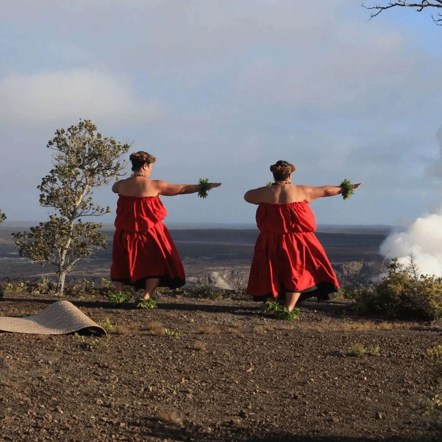 Two women in red traditional dresses performing a dance outdoors with volcanic landscape and smoke in the background.