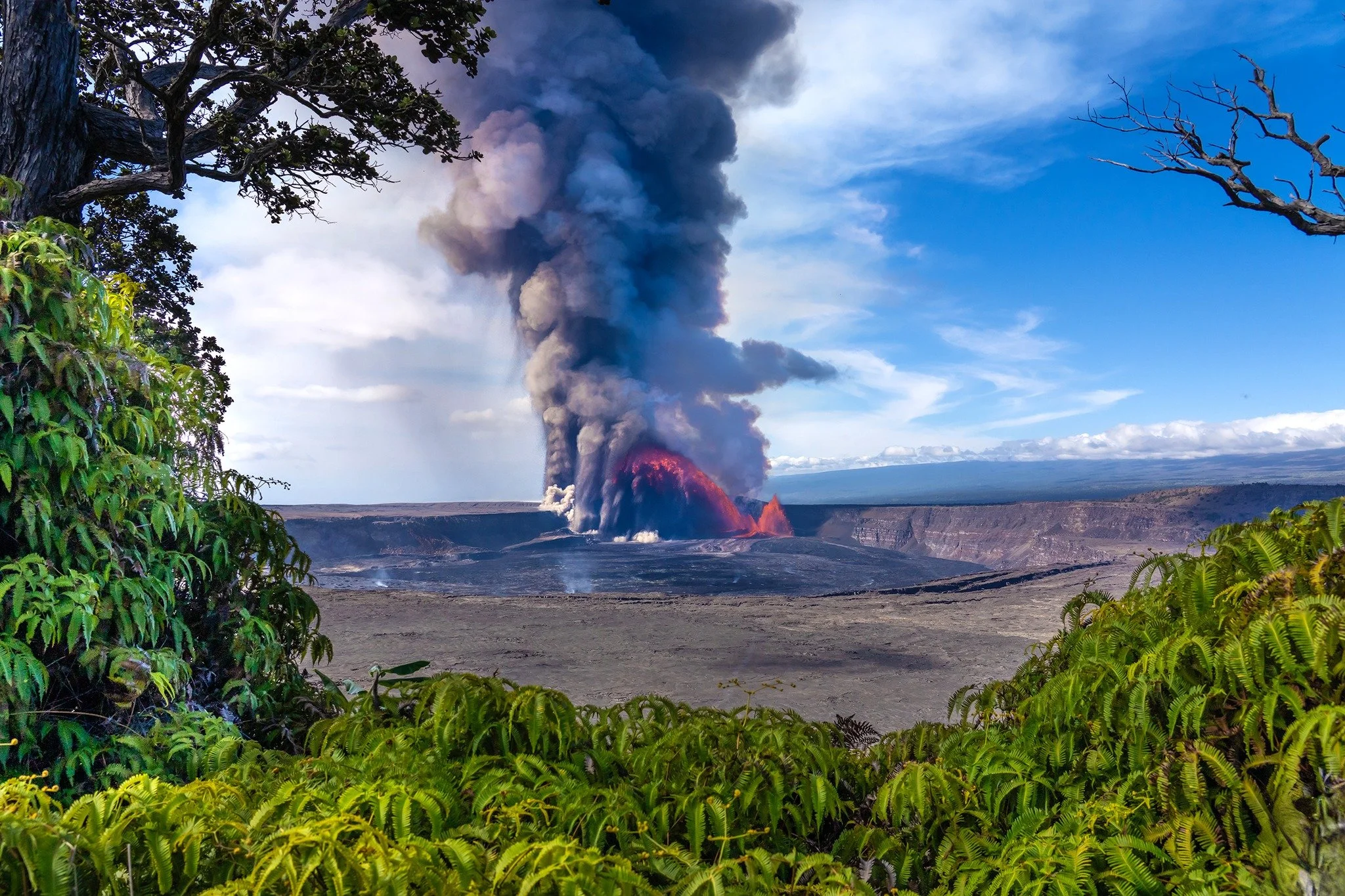A volcanic eruption with red lava flowing and dark smoke billowing into the sky, framed by green foliage and trees in the foreground, under a partly cloudy sky.
