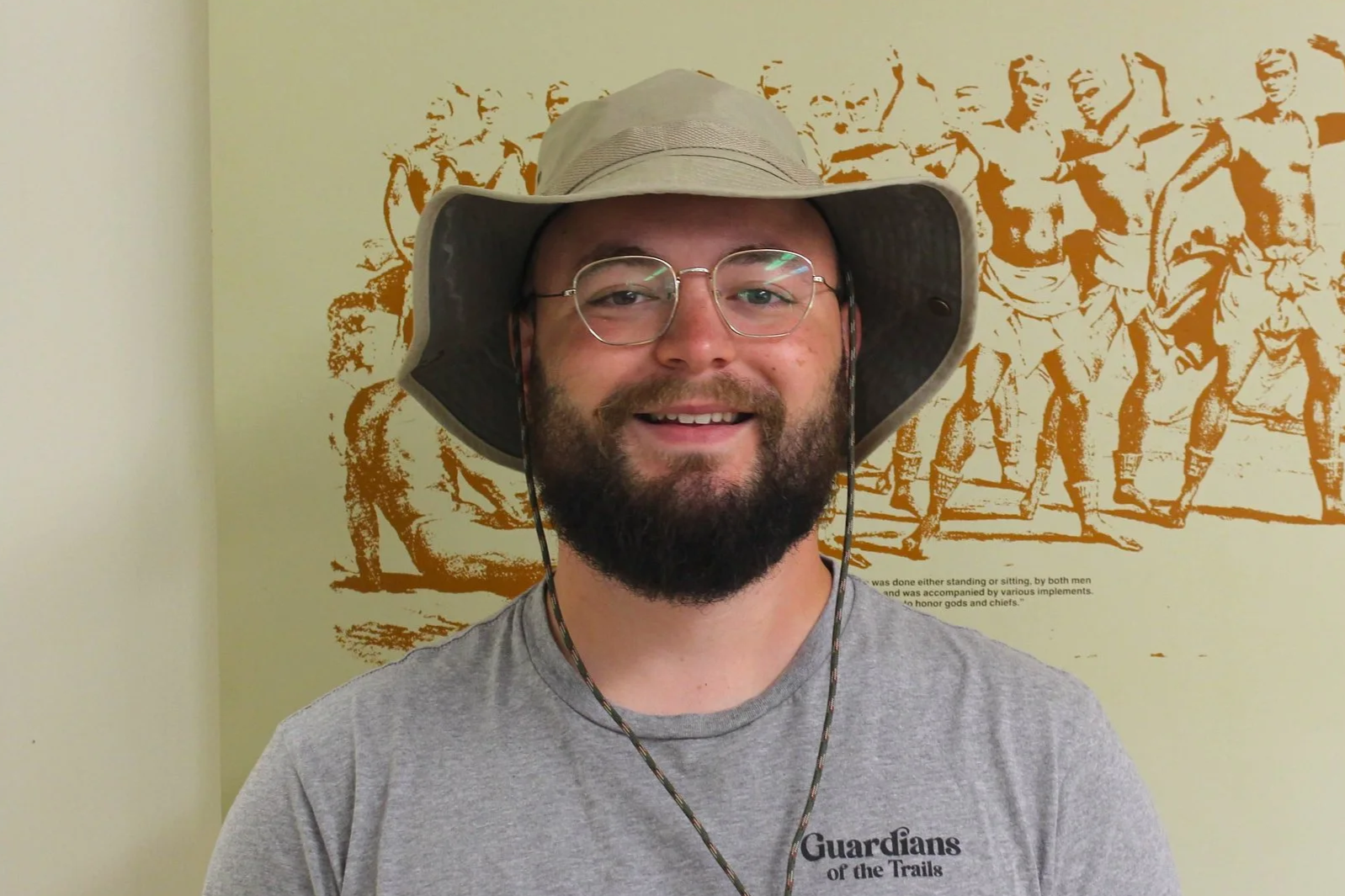 A man wearing glasses, a gray hat, a gray t-shirt with 'Guardians of the Trails' written on it, and a beard, smiling at the camera. Behind him is a colorful wall art depicting multiple figures in a vintage style.