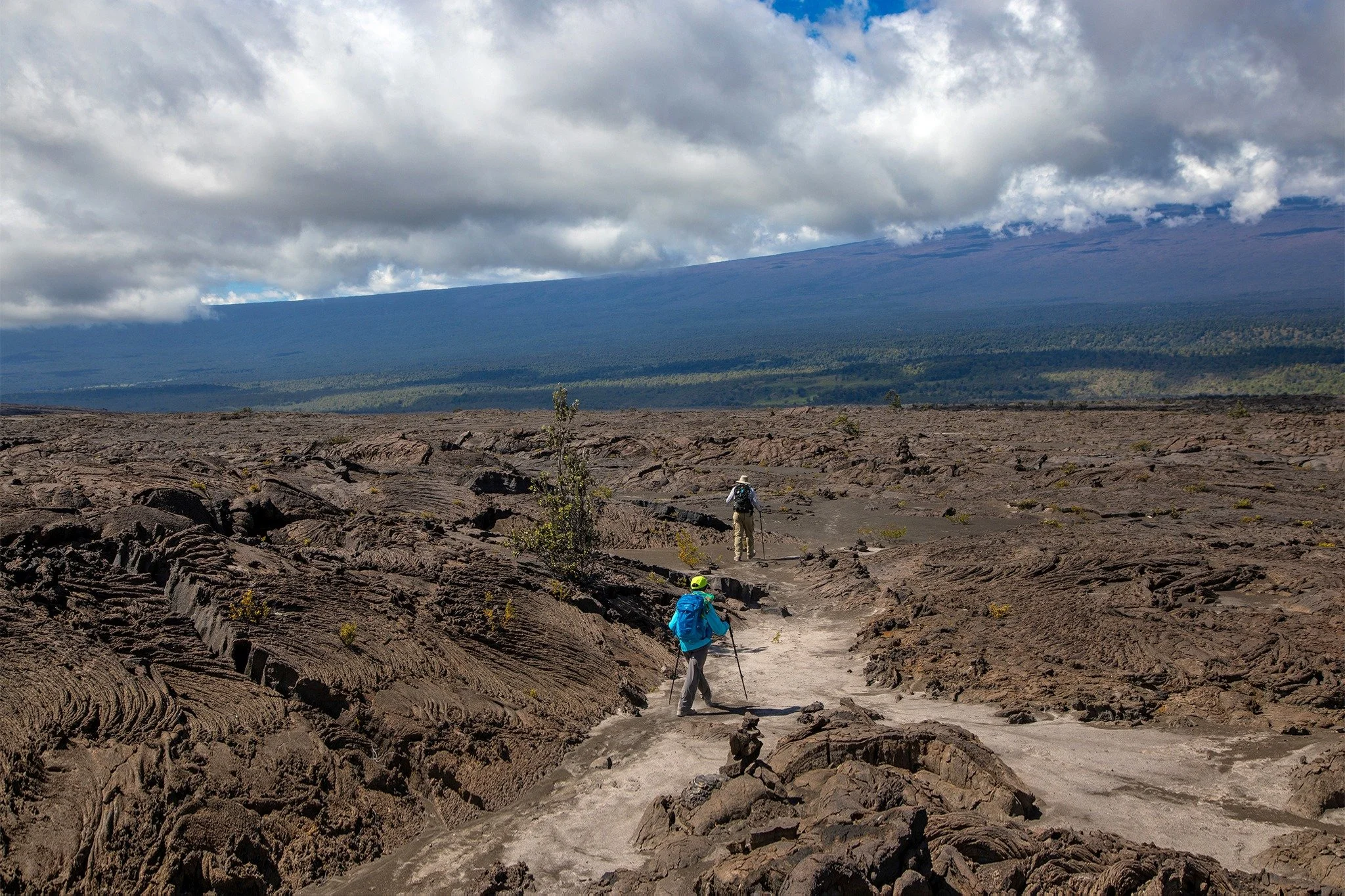 Hikers walking on a rocky volcanic terrain under a cloudy sky with large clouds and patches of blue sky, a mountain in the background, and some sparse vegetation.