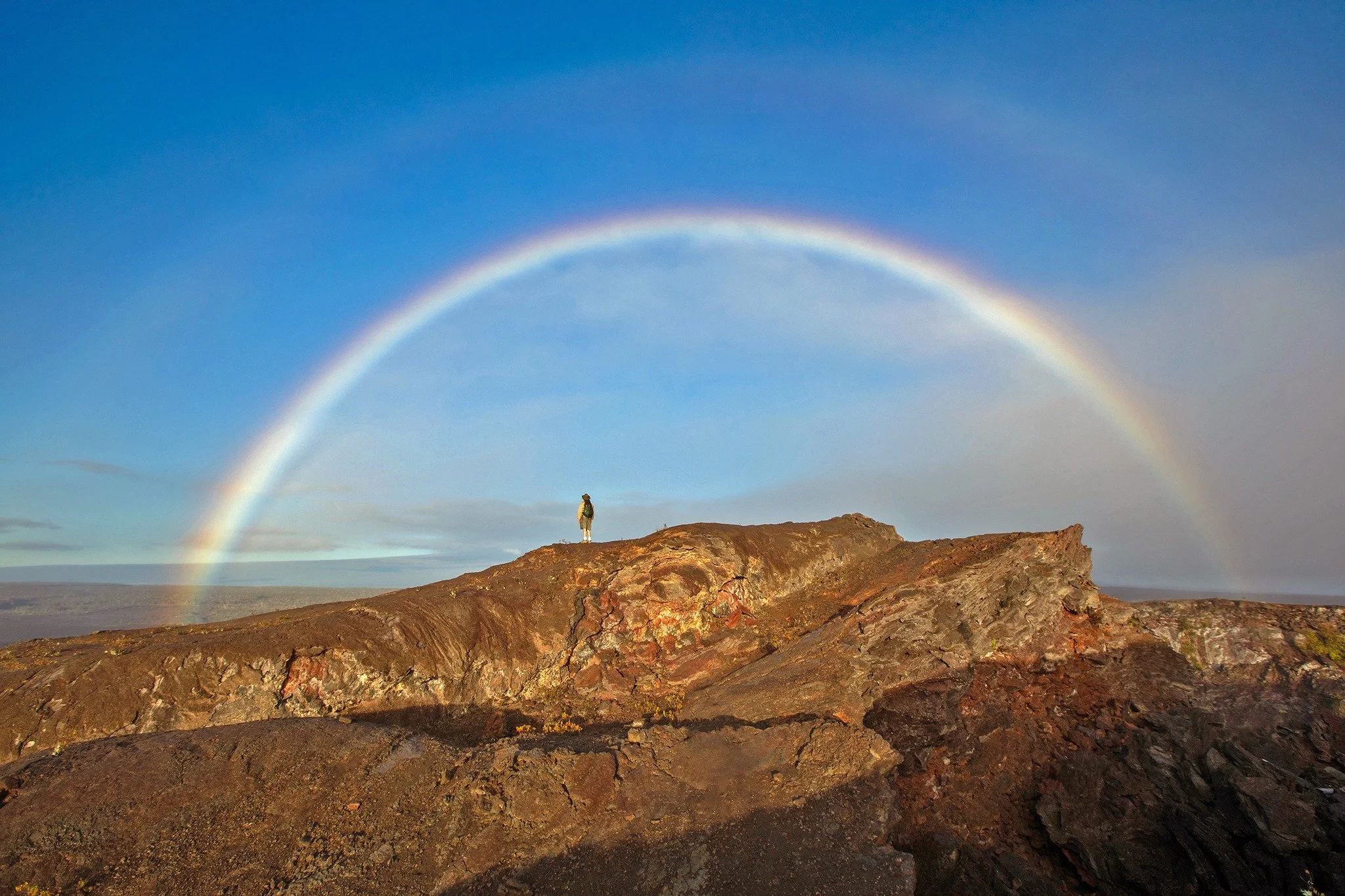 A person standing on rocky terrain under a rainbow in the sky.