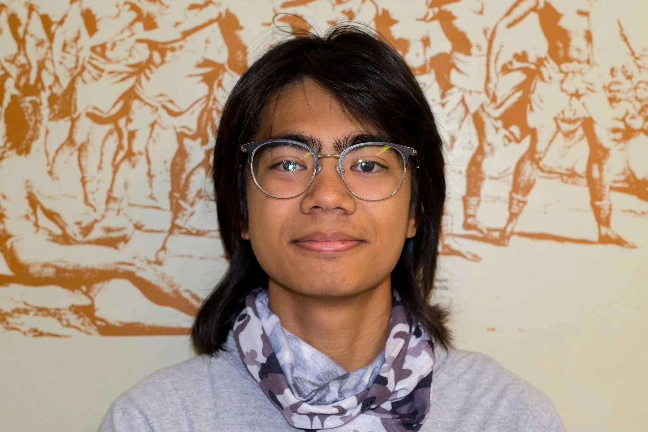 A person with short dark hair, glasses, and a patterned scarf, standing in front of a wall with orange abstract artwork.