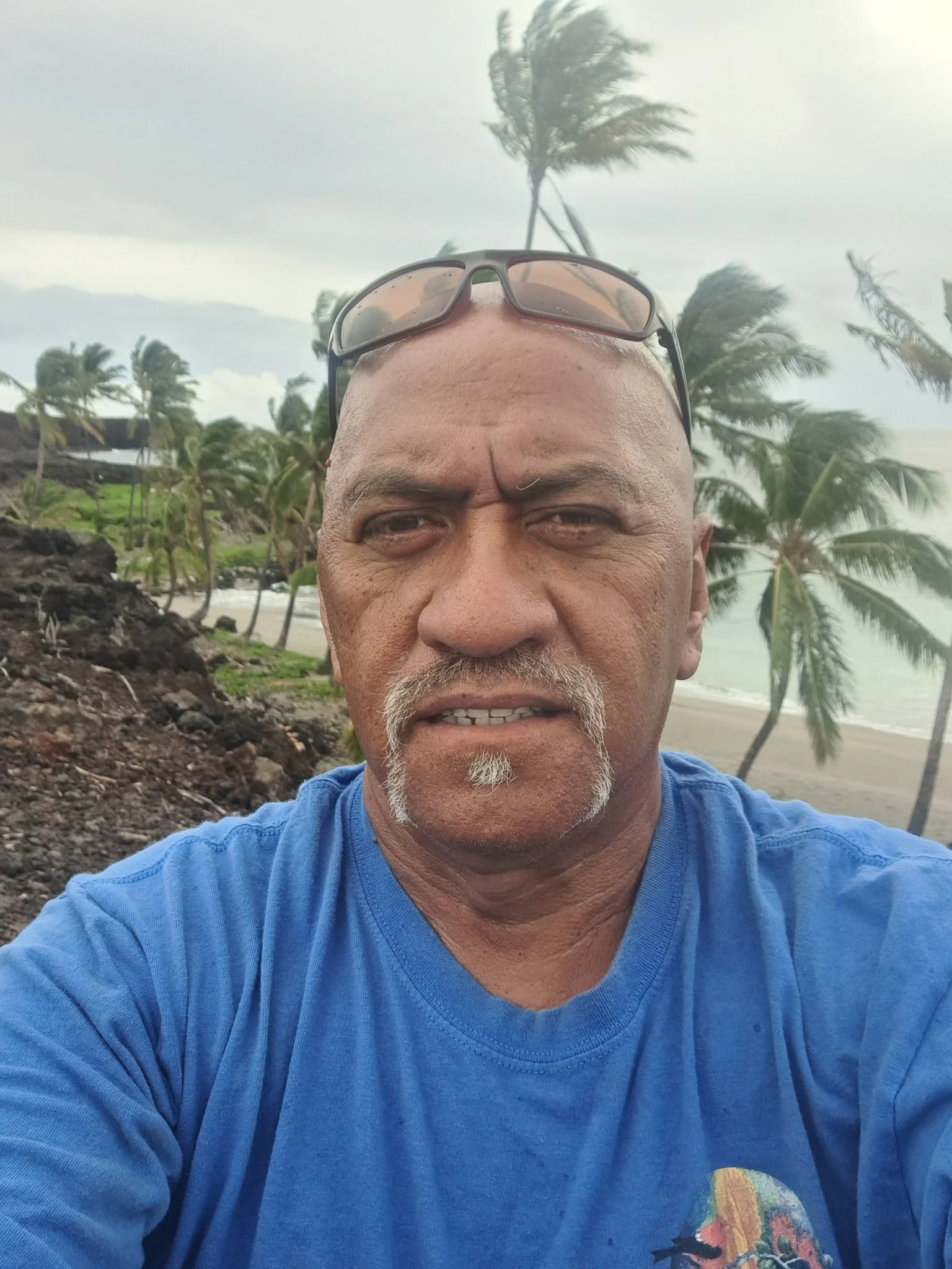 A man with a shaved head and sunglasses on top of his head taking a selfie on a tropical beach with palm trees blowing in the wind and cloudy skies in the background.