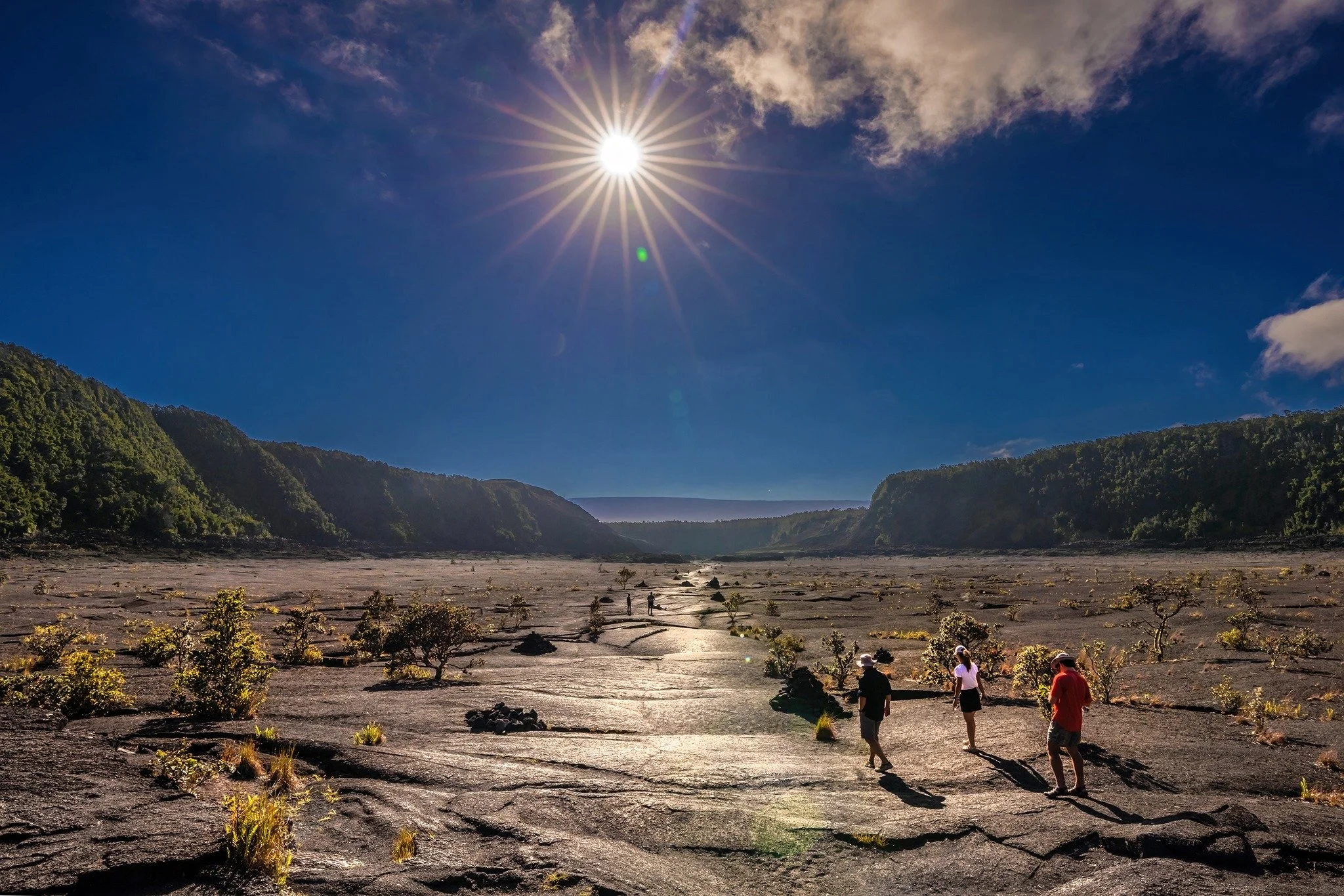 Group of people walking on a dry riverbed with small plants, surrounded by mountains under a bright sun in a clear blue sky with some clouds.