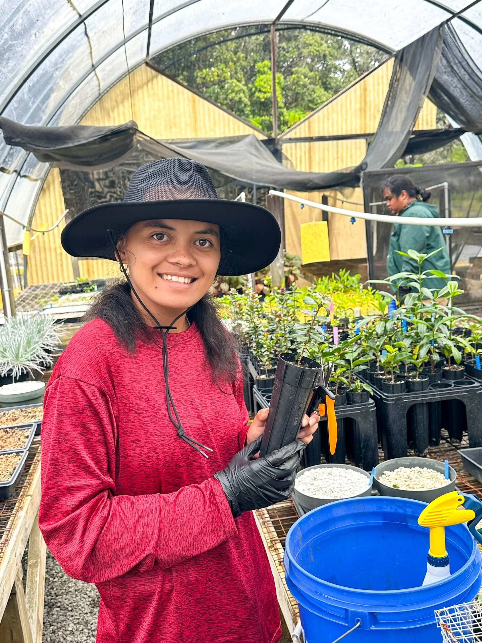A woman in a red long-sleeve shirt and black wide-brimmed hat is smiling and holding gardening tools inside a greenhouse. In the background, there are two other people and several potted plants, with gardening supplies and watering tools nearby.