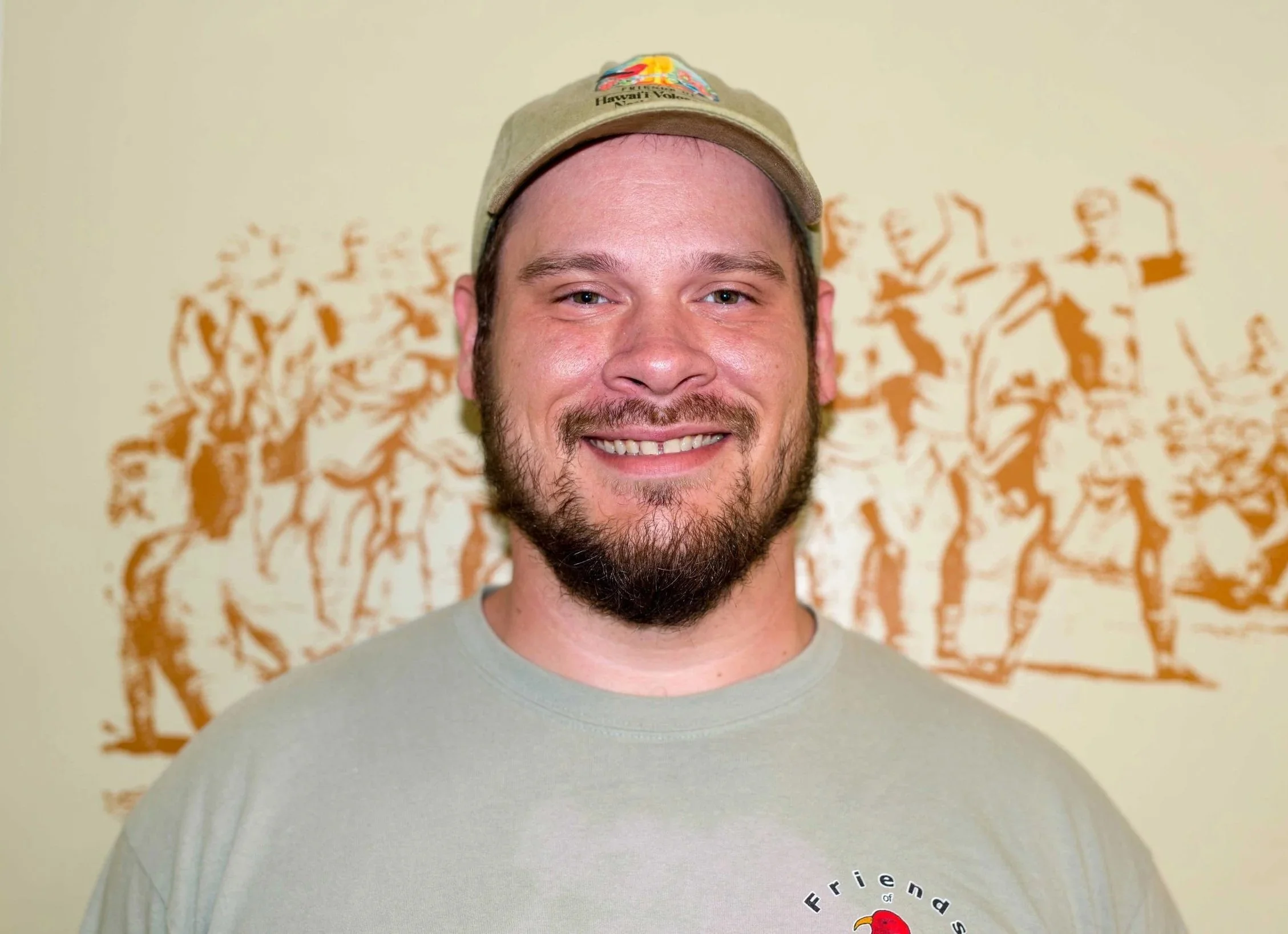 A smiling man with a beard and mustache wearing a beige cap with a colorful patch and a gray T-shirt, standing in front of a wall decorated with orange and brown animal silhouettes.