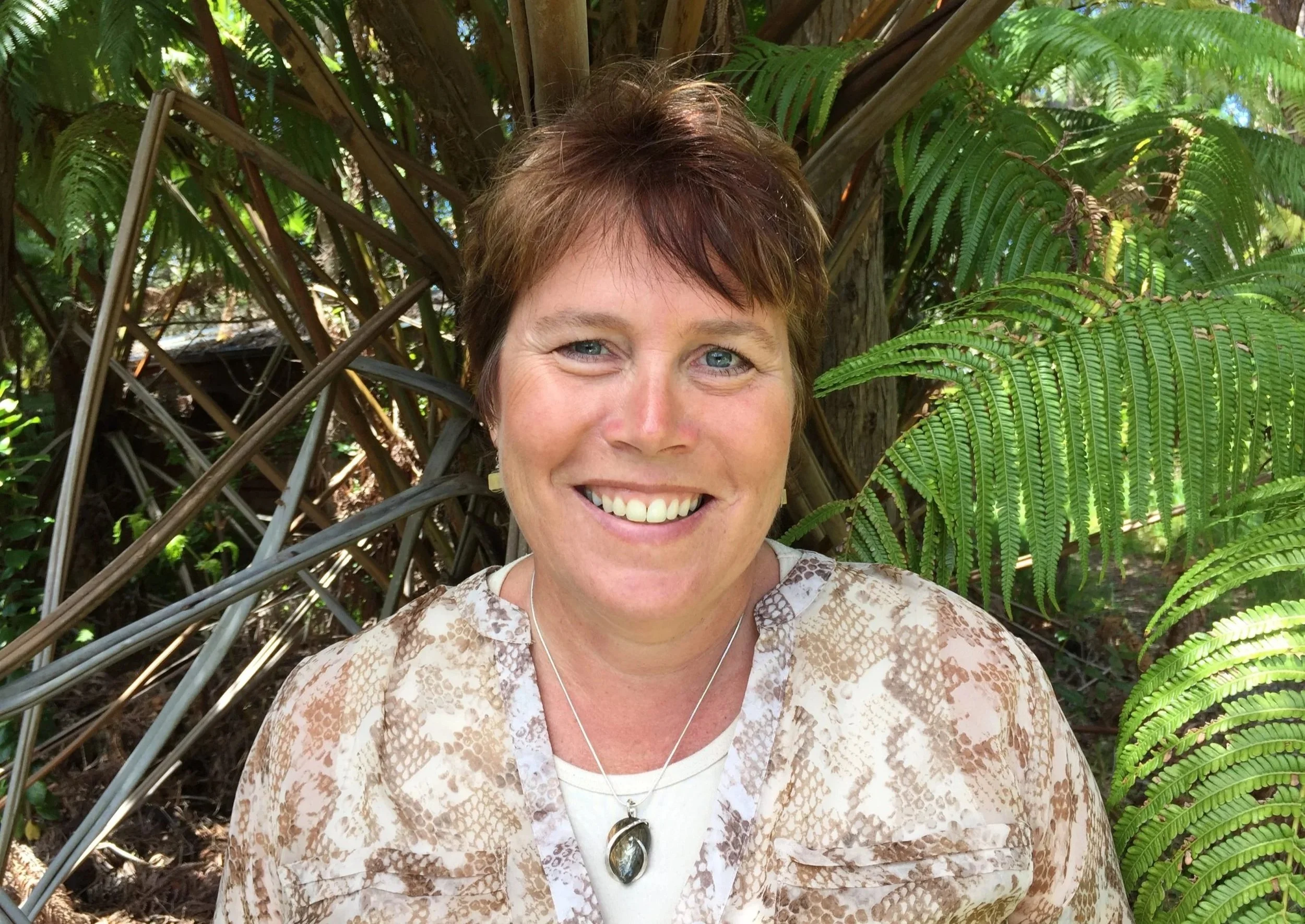 A woman with short brown hair and blue eyes smiling outdoors, standing among lush green ferns and tropical plants.