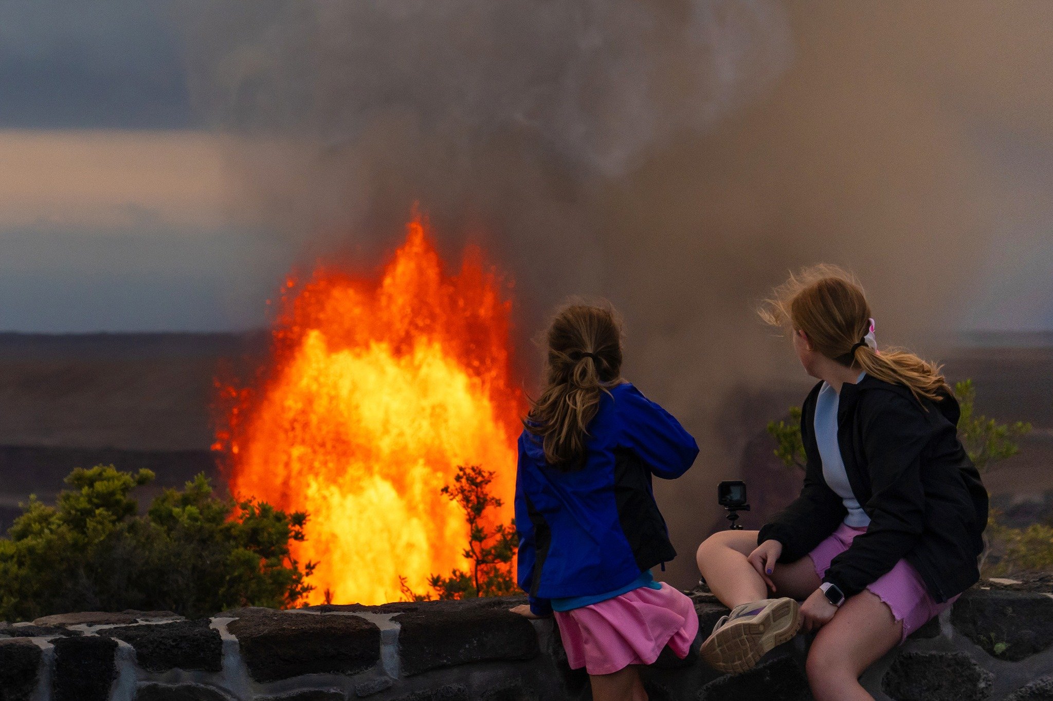 Two children sitting on a stone wall watching a large eruption of lava and ash from a volcano in the distance during dusk, with dark clouds overhead.