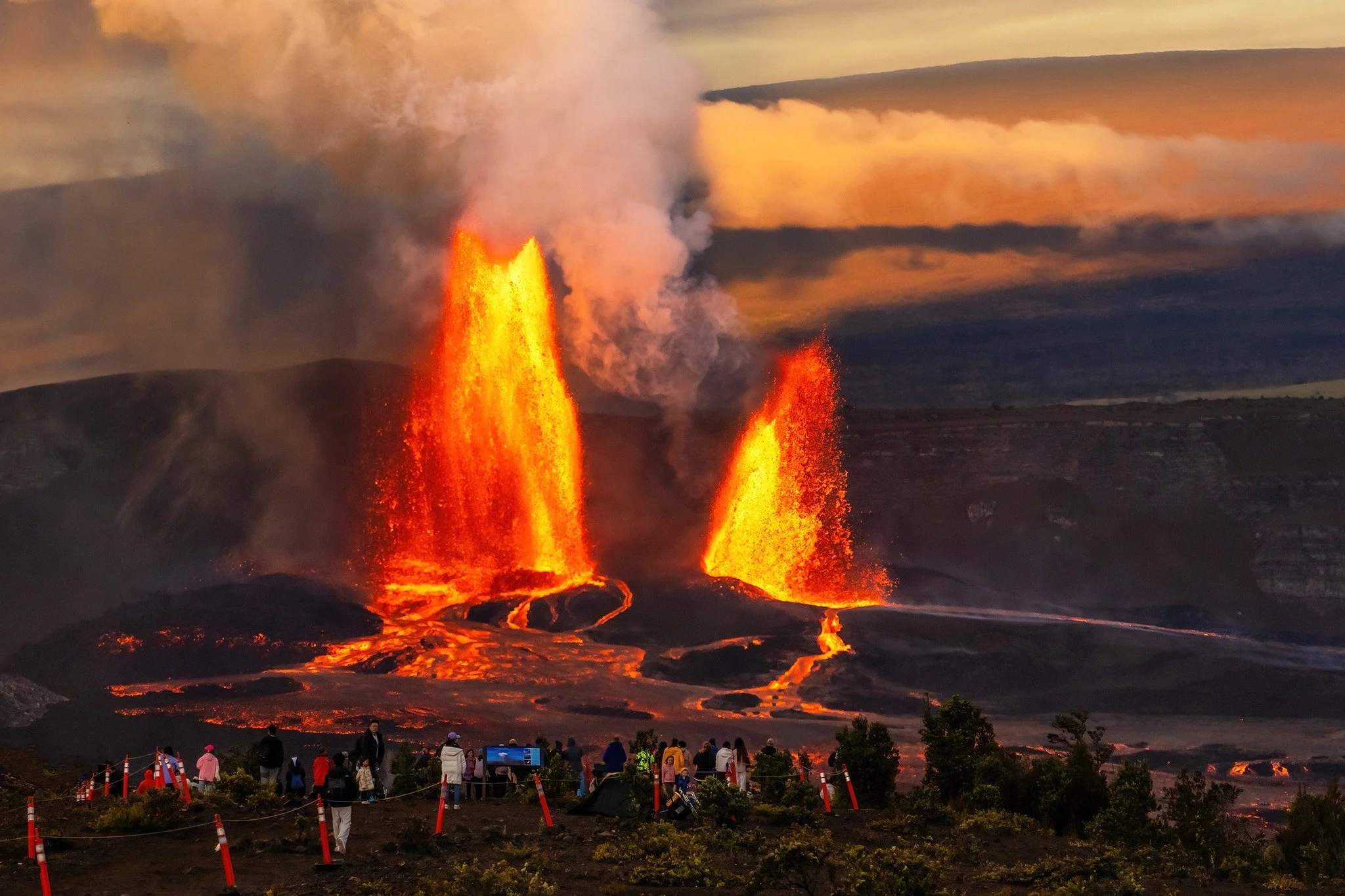 A volcanic eruption with lava and smoke rising from the volcano, with a group of spectators and tourists observing from a designated viewing area in the foreground.