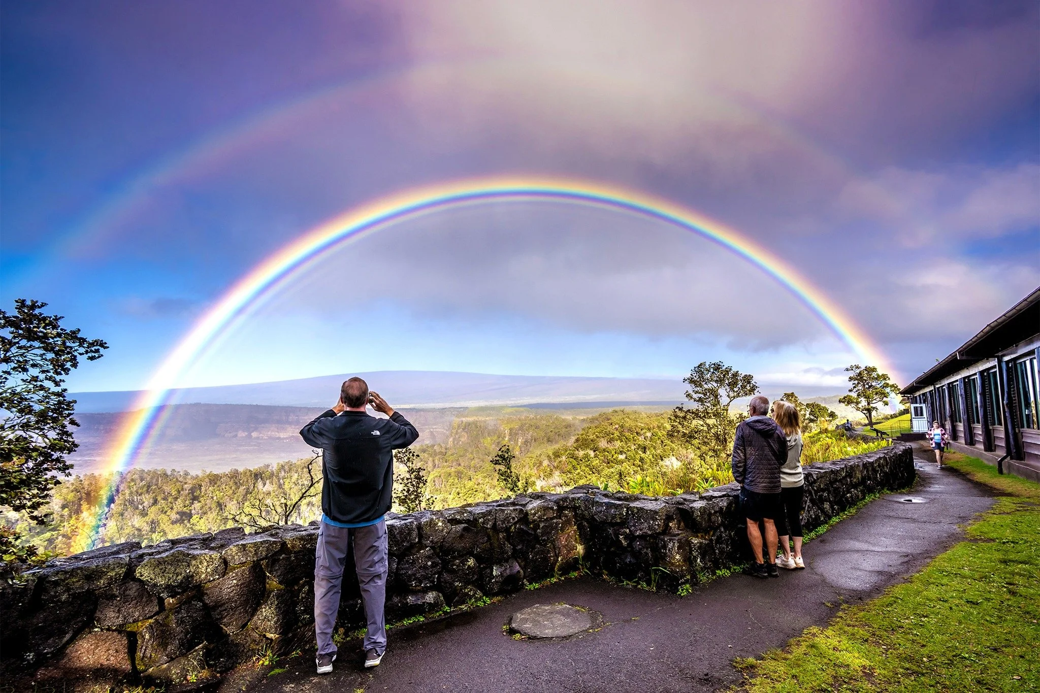 A view of a double rainbow in the sky over a scenic landscape with trees, hills, and a cloudy sky, as people observe and photograph the scene from a stone wall and pathway.