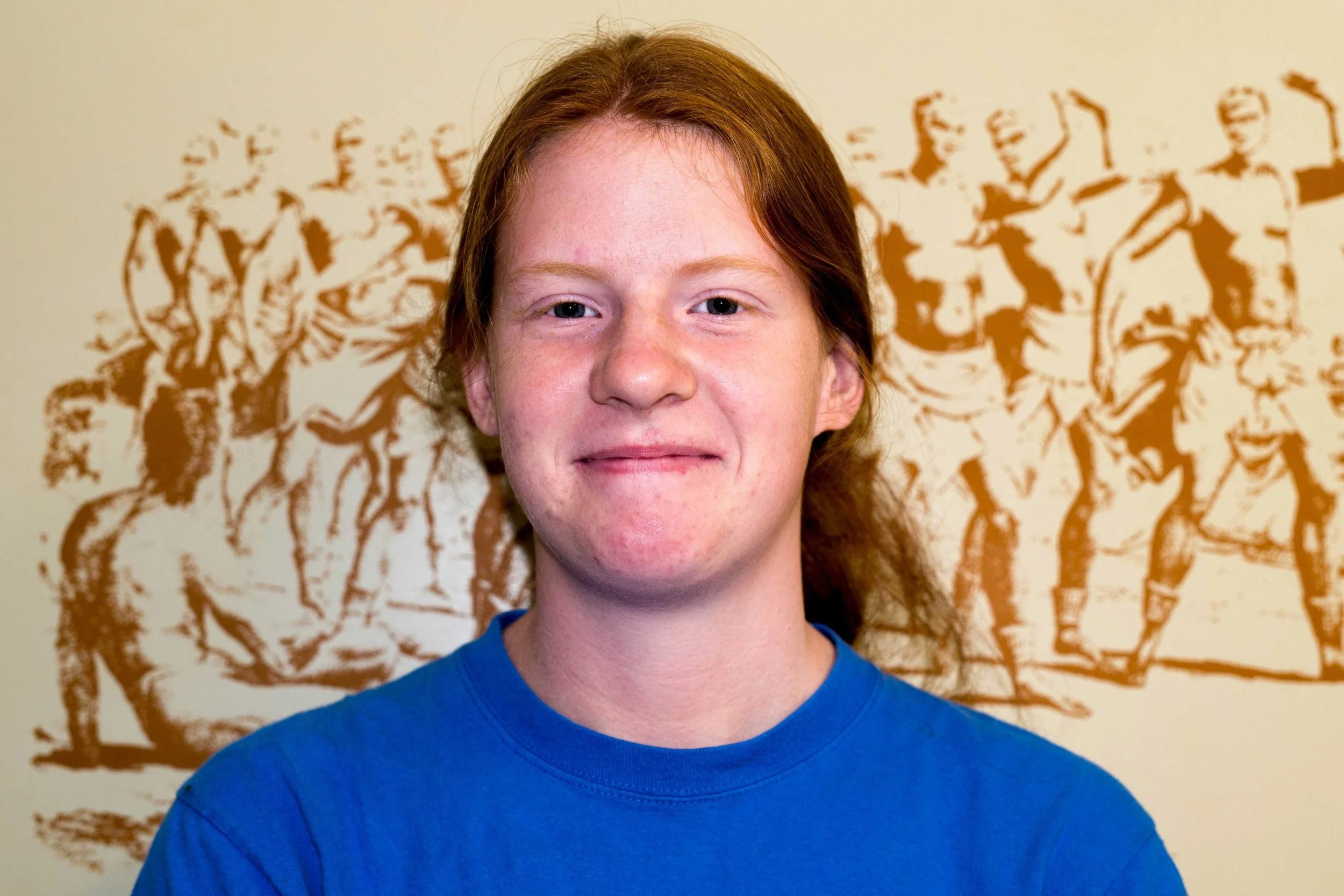 Portrait of a young person with red hair, wearing a blue shirt, standing in front of a wall with a stencil art of soldiers in army uniforms.