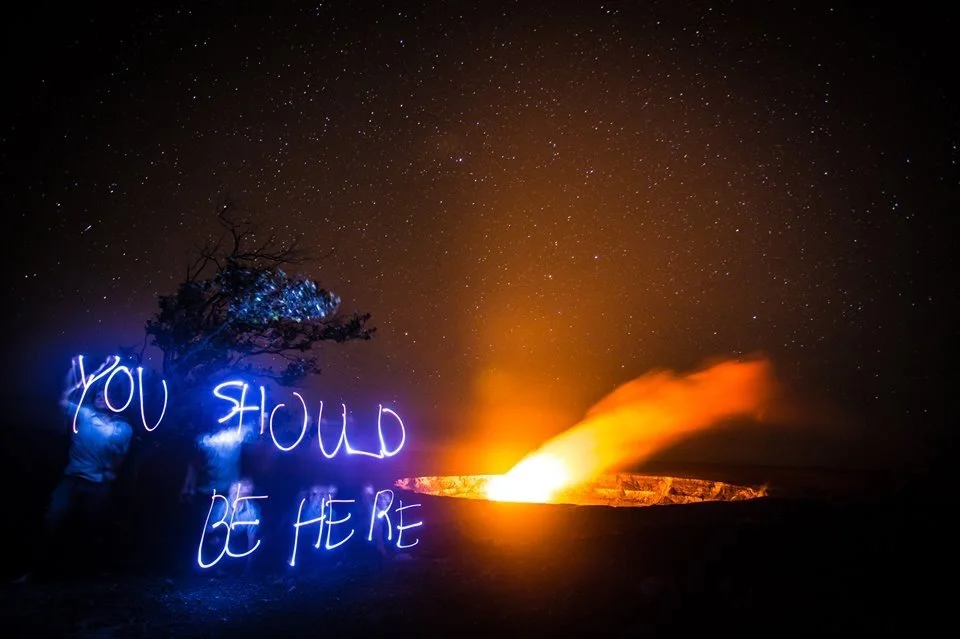 A person near a tree writes 'YOU SHOULD BE HERE' with a blue light in the darkness under a starry sky, with a volcano erupting in the background, emitting bright orange lava and smoke.