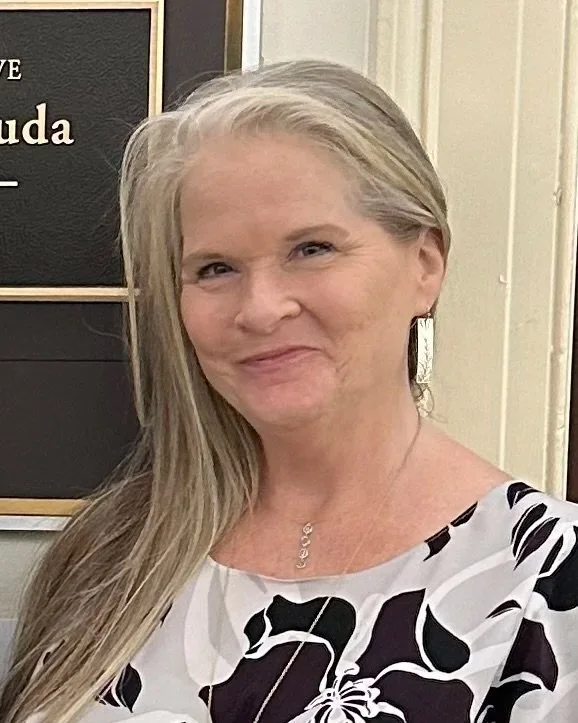 A smiling middle-aged woman with long blonde hair, wearing a white blouse with black floral patterns, standing indoors near a blackboard with text, in a well-lit room.