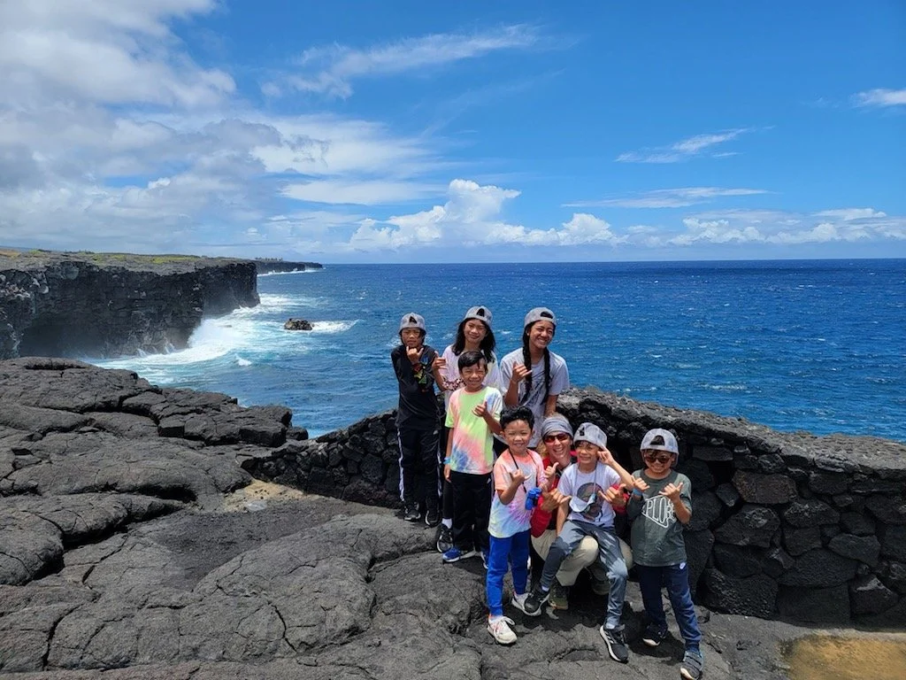 Group of children and adults posing on black volcanic rocks near the ocean with cliffs and blue sky with clouds in the background.