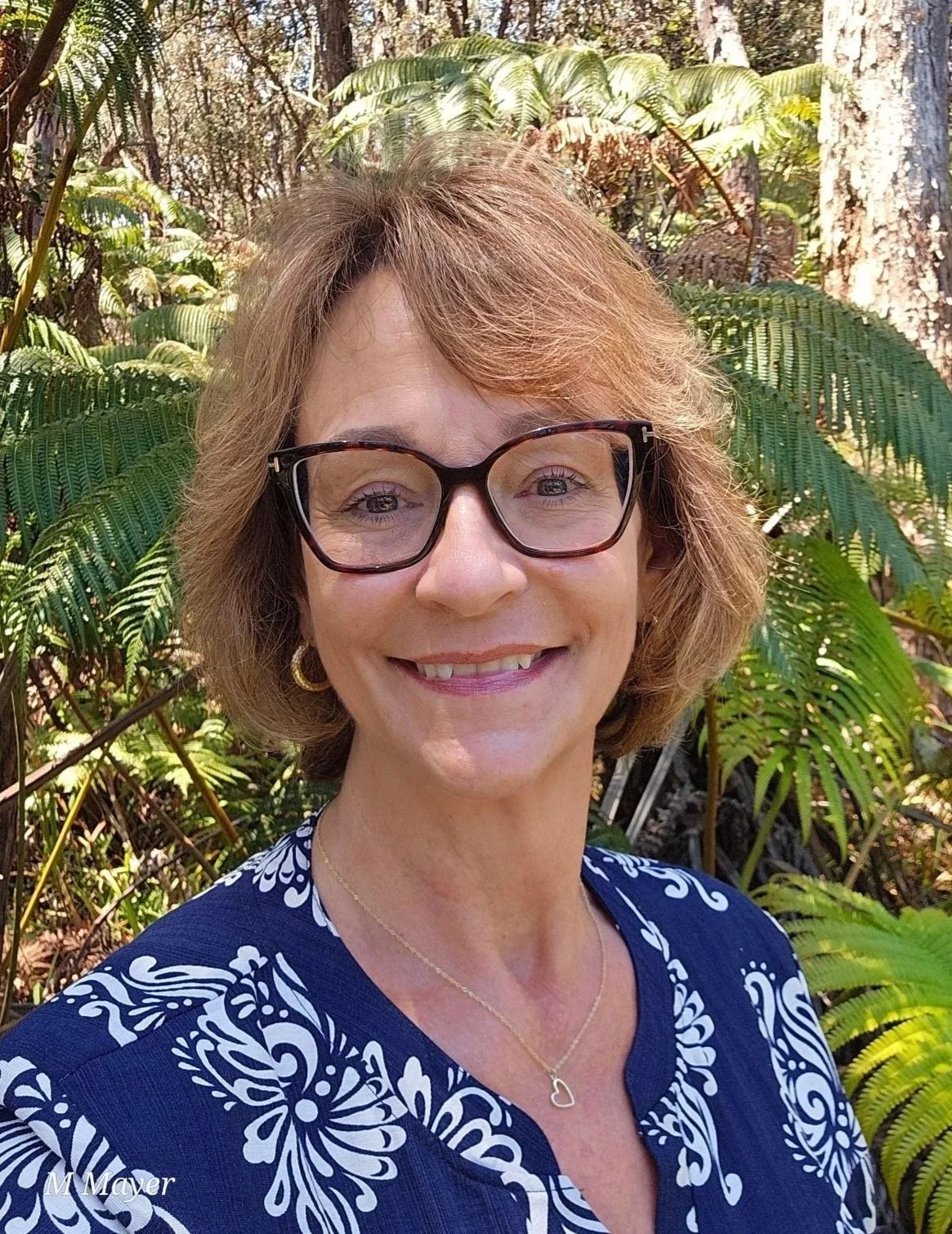 A woman smiling outdoors in front of green fern plants, wearing glasses, a blue and white patterned top, gold hoop earrings, and a delicate heart necklace.
