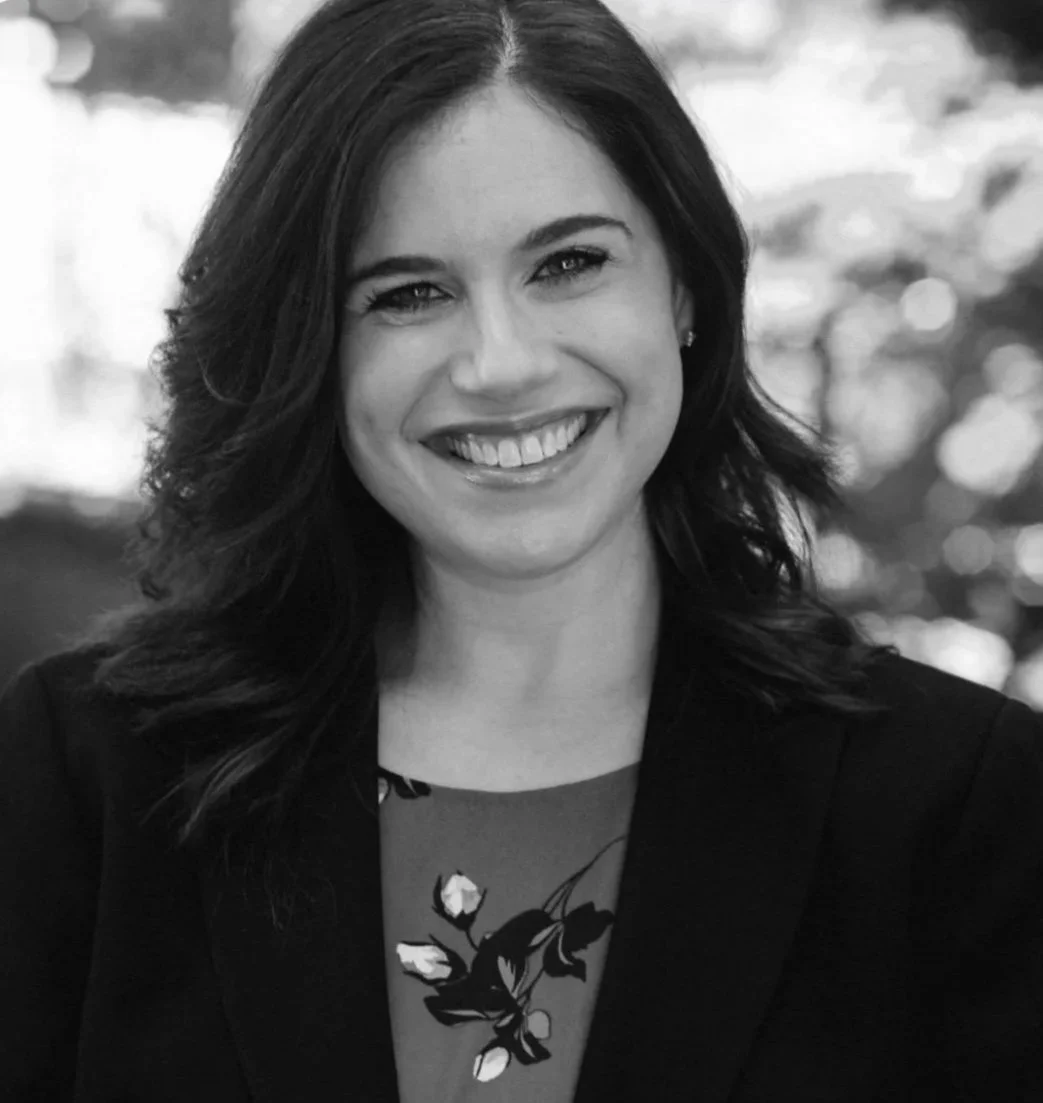 Black and white portrait of a woman smiling outdoors, with shoulder-length wavy hair, wearing a dark blazer and a floral-patterned top.