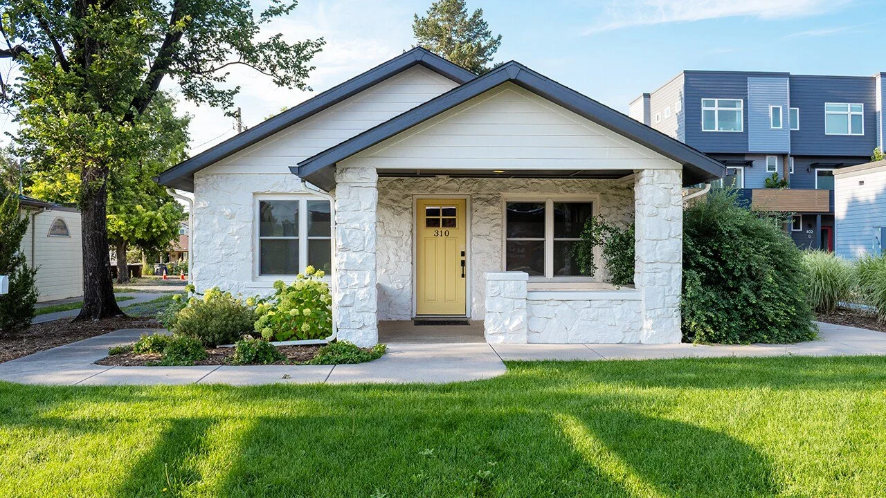 White house with stone exterior, yellow front door, surrounded by green lawn and trees, with neighboring modern buildings in the background.