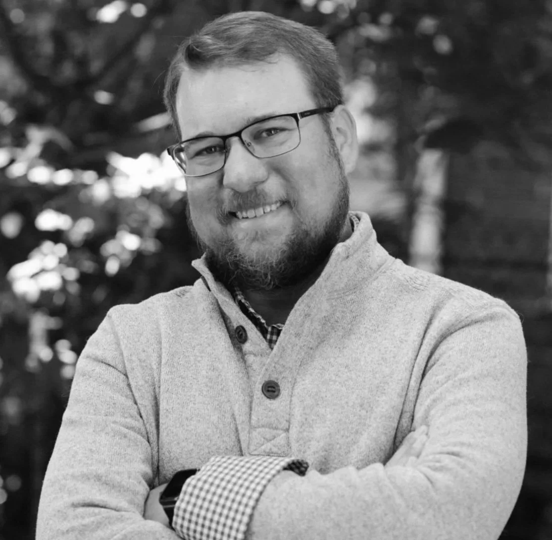 Black and white portrait of a man with glasses, a beard, and a smiling expression, standing outdoors with blurred trees in the background.