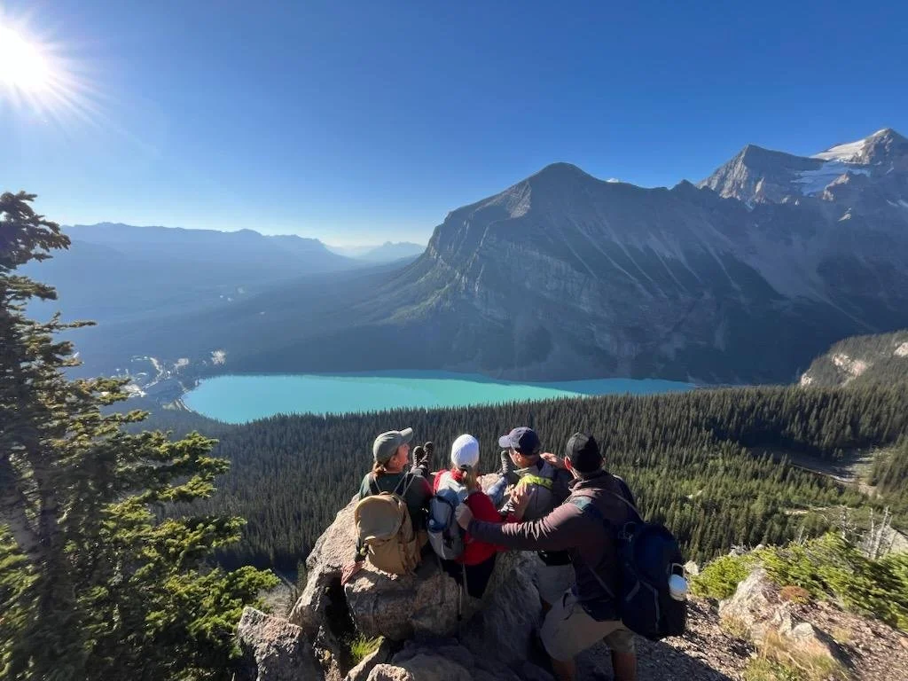 Group of six hikers resting on a rocky ledge overlooking a turquoise lake surrounded by dense forest and towering snow-capped mountains under a clear blue sky.