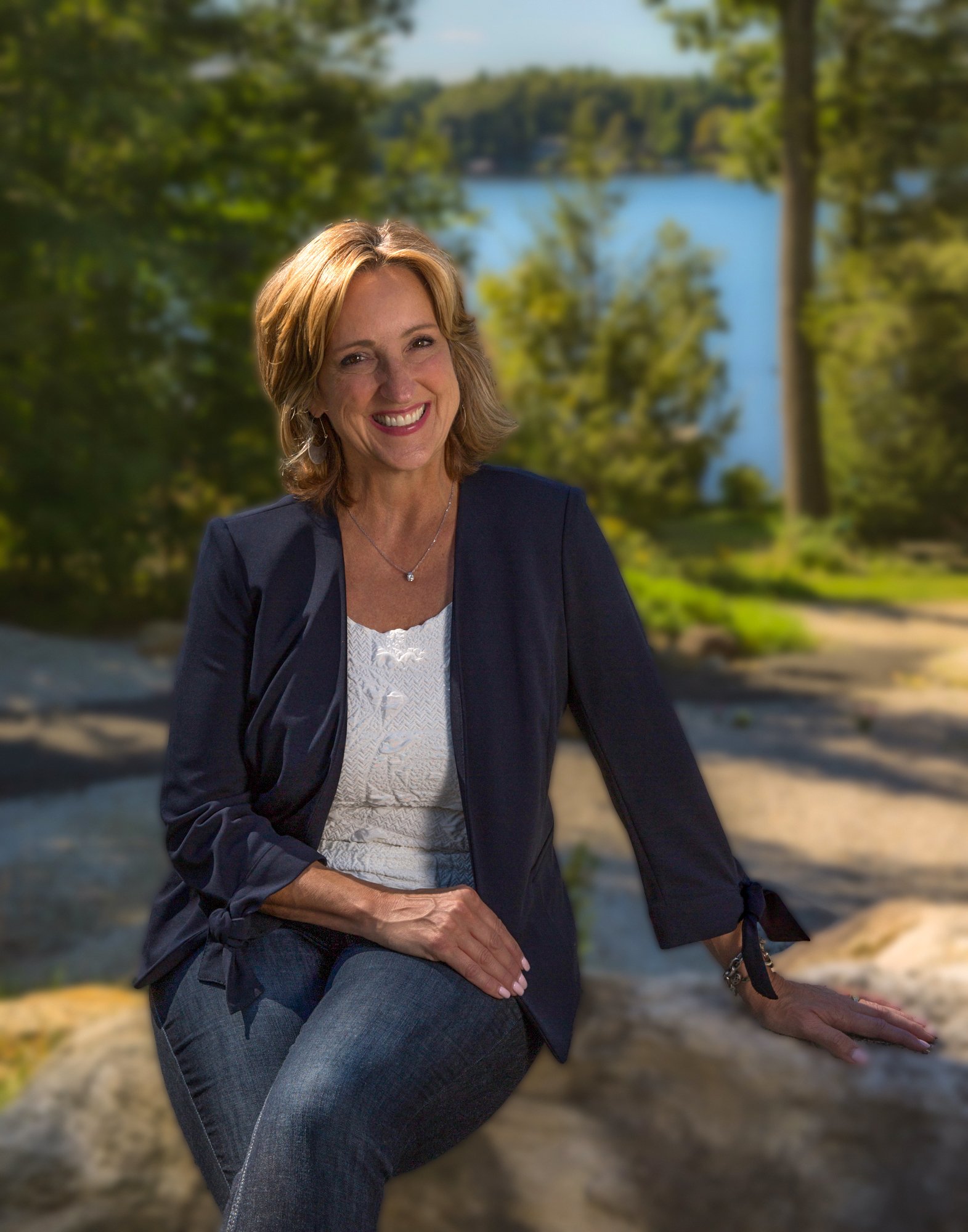 Chris Chaia sitting on a large rock outdoors with trees and a lake in the background, smiling at the camera.