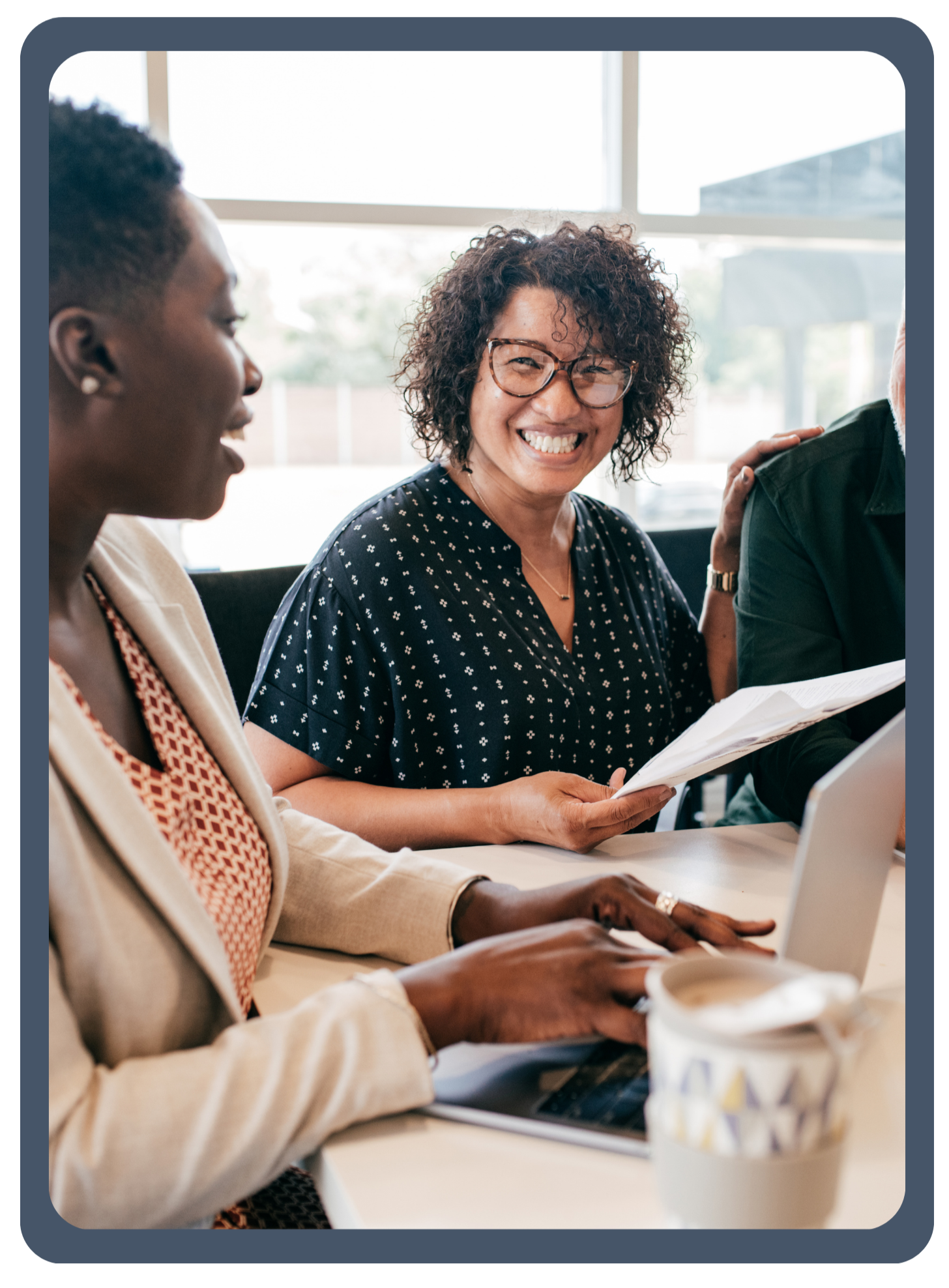 Two women are sitting at a table in a bright office, smiling and engaged in a discussion, with laptops and documents in front of them.