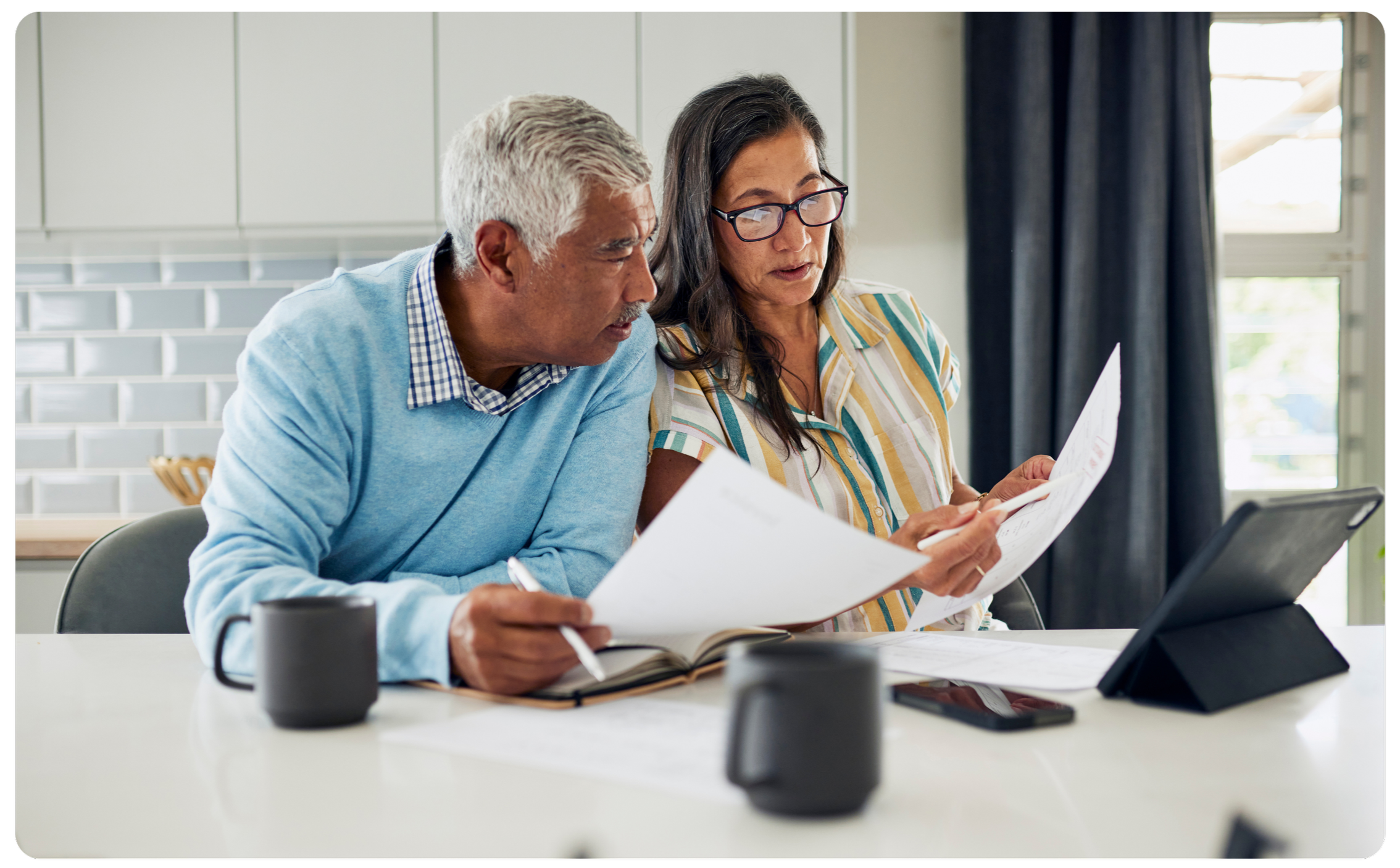An elderly couple sitting at a table, reviewing documents and looking at an electronic device, in a room with a window and curtains.