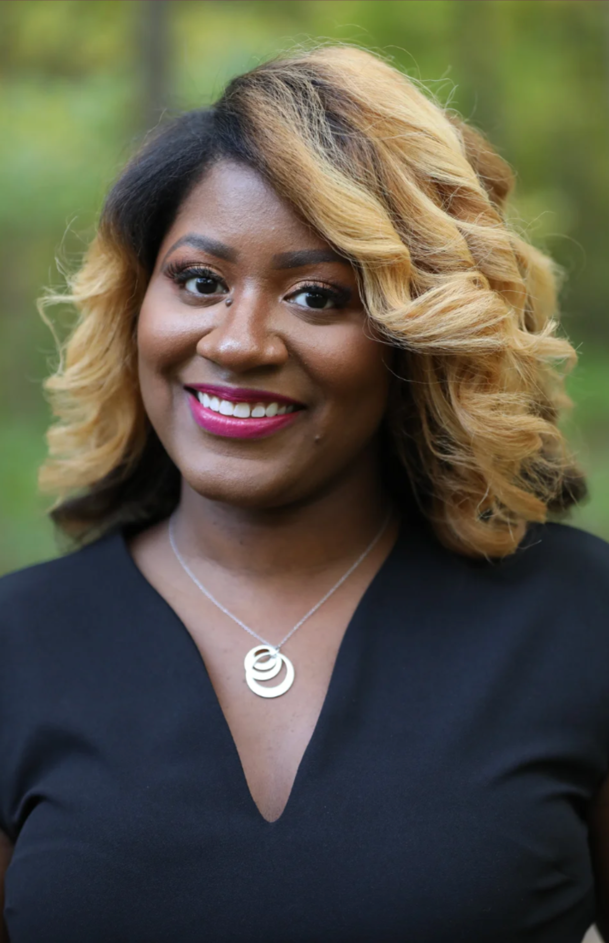 A woman with shoulder-length, curly blond hair and dark roots, smiling outdoors with a blurred green background. She wears a black top and a silver necklace.