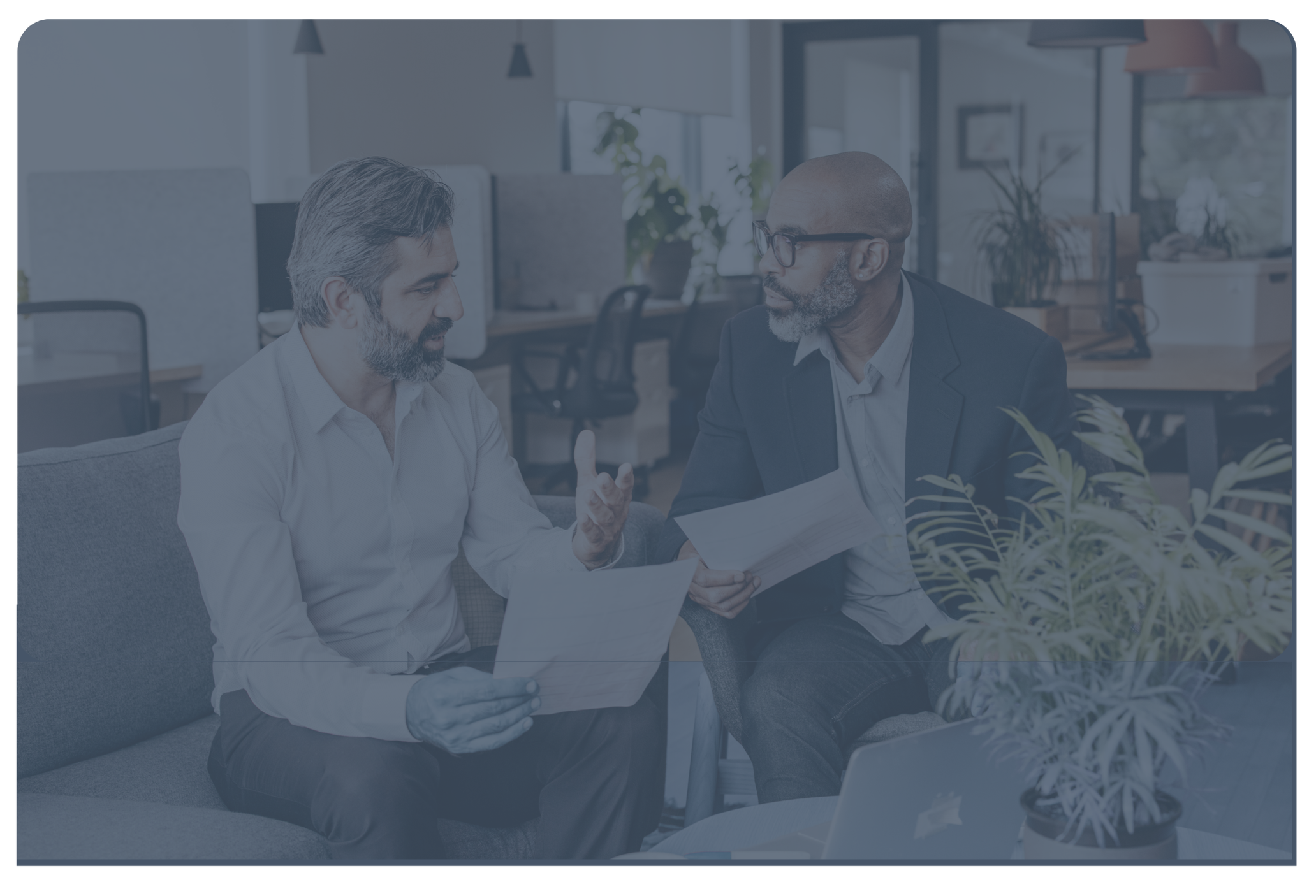 Two men having a discussion in an office. One man has gray hair and a beard, and the other is bald with glasses. They are sitting on a couch, holding papers, and gesturing while talking.