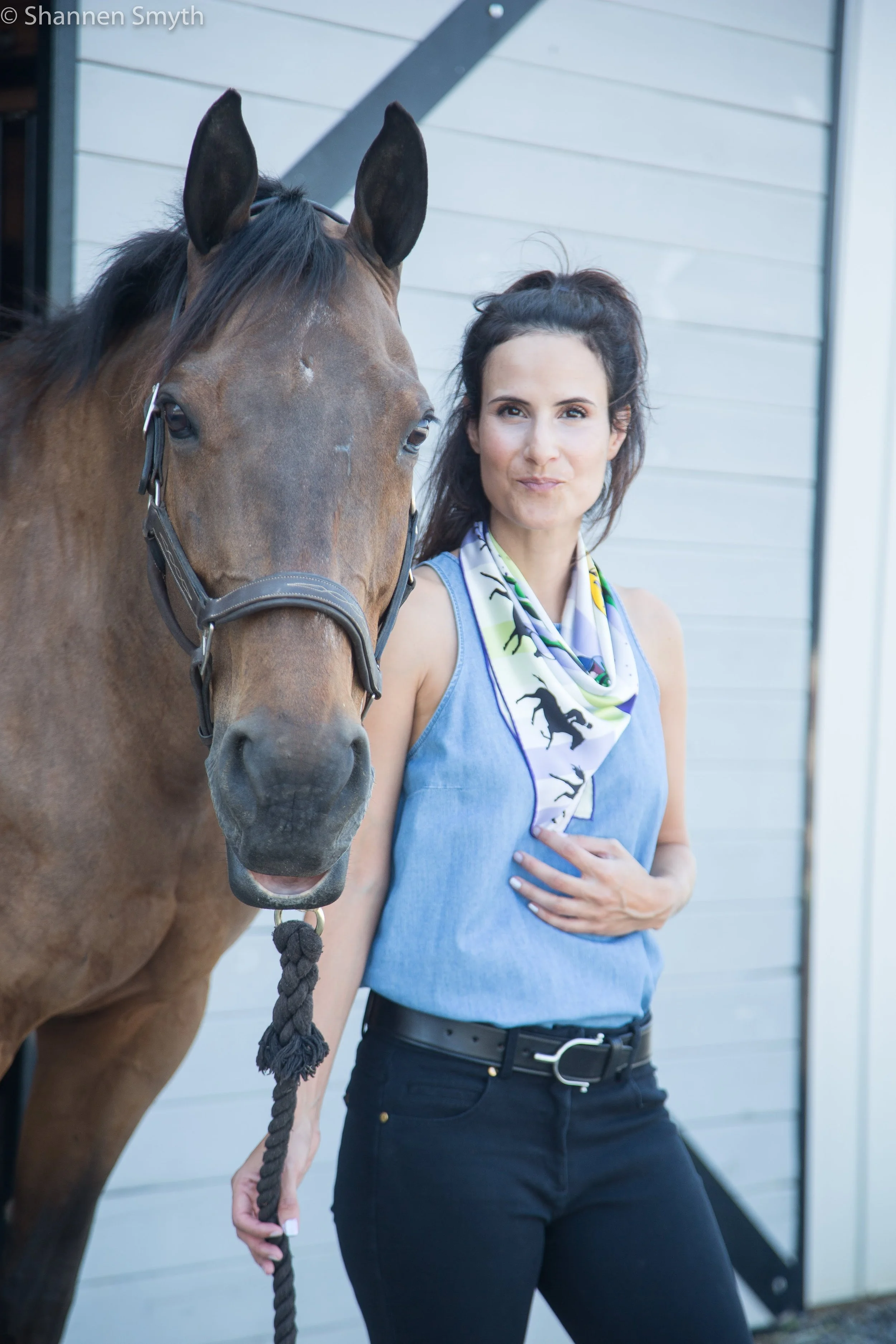 A woman standing next to a brown horse, touching her chest with her left hand, in front of a light-colored barn with gray siding.
