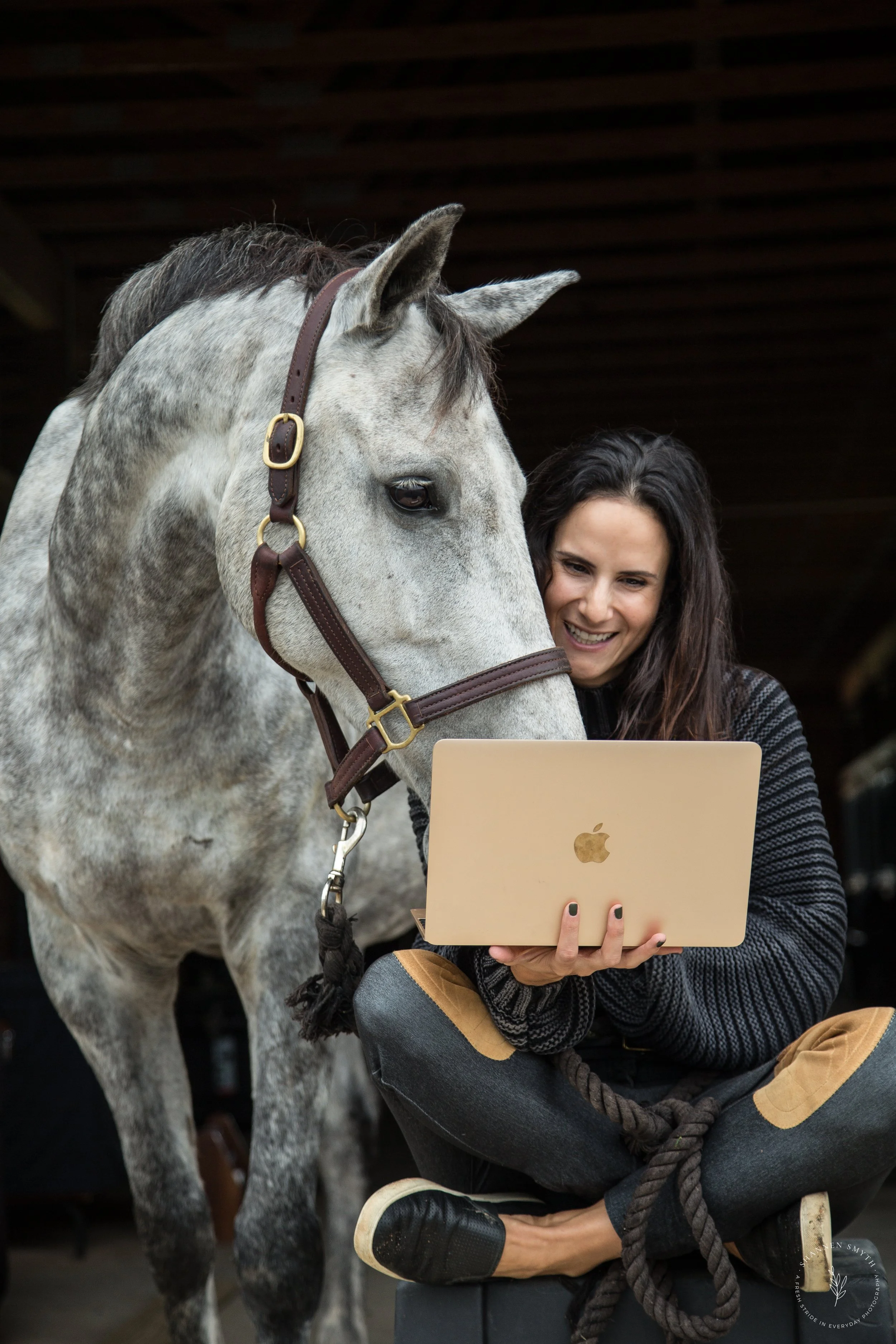 A woman sitting cross-legged on a black box, smiling and looking at a gold MacBook, while a gray horse with a dark mane stands close, also looking at the laptop.