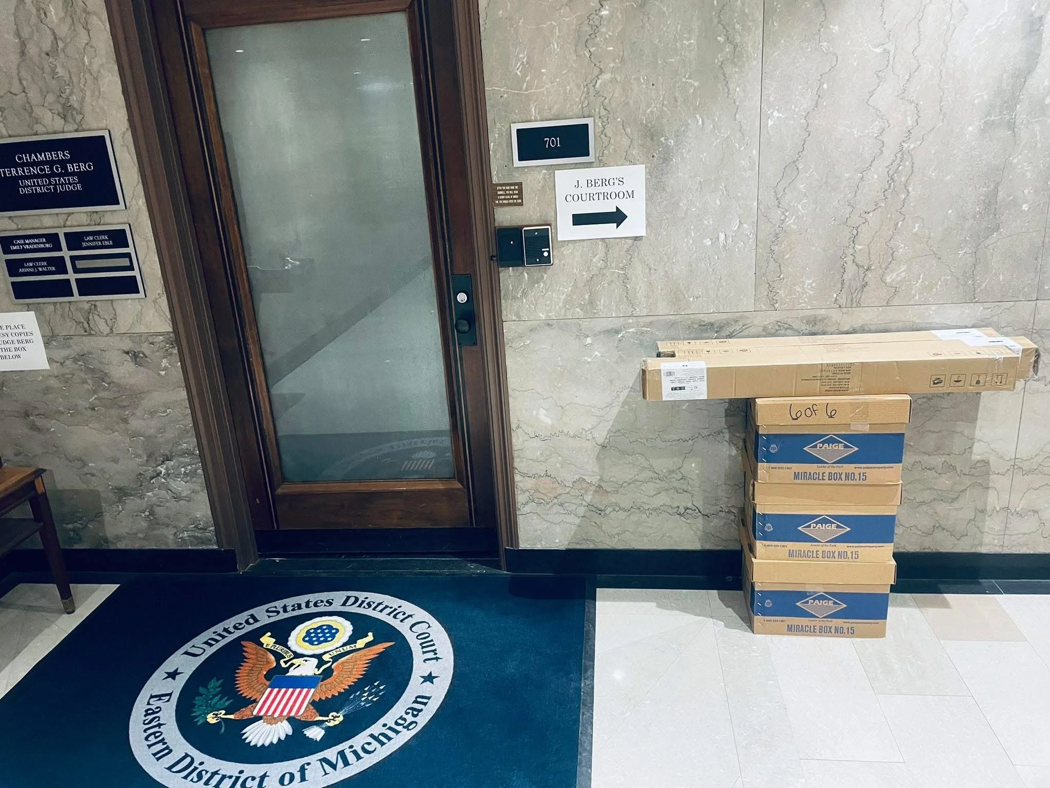 The image shows a hallway with beige marble walls and floors. Near the center, there's an elevator door with a blue panel displaying the number 701. To the right of the elevator, a sign pointing to the right reads "J. Berge's Courtroom." To the left of the elevator, there are several blue nameplates on the wall with various titles and names, including a reference to a United States District Judge. At the bottom left, part of a round floor emblem with the United States District Court seal is visible. To the right, there are multiple stacked cardboard boxes labeled "Miracle Box No. 15" and