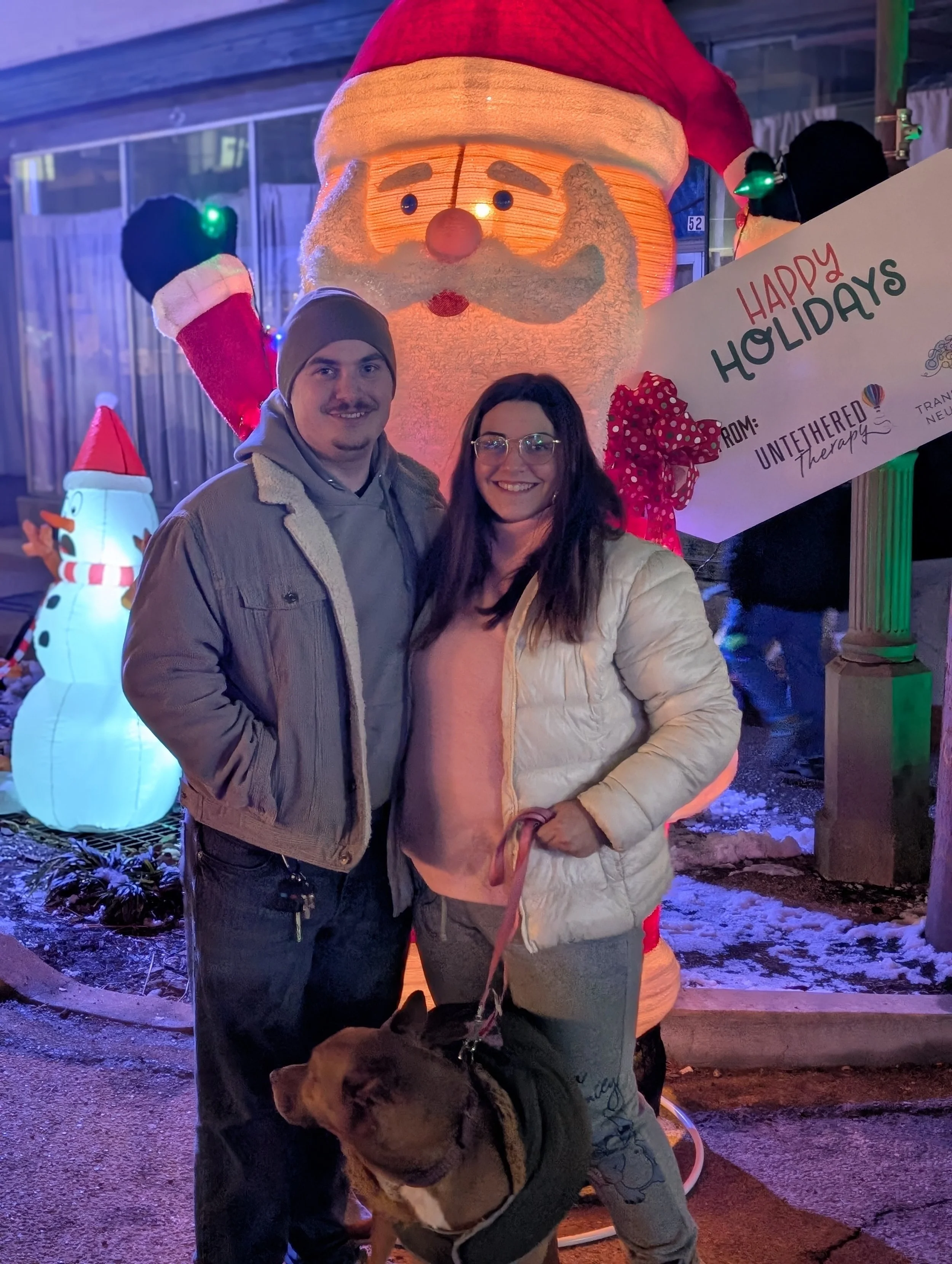 A man and woman standing outside at night, smiling at the camera. Behind them are large holiday decorations including a giant Santa Claus and a snowman, with a sign that reads 'Happy Holidays from: Untethered Therapy.' The woman is holding a dog on a leash.