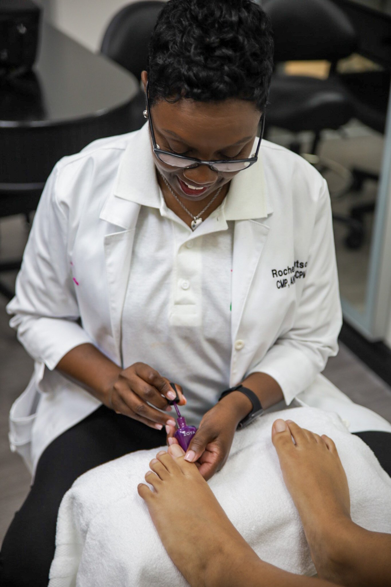 A woman in a white lab coat applying purple nail polish to a person's toenails during a pedicure treatment.