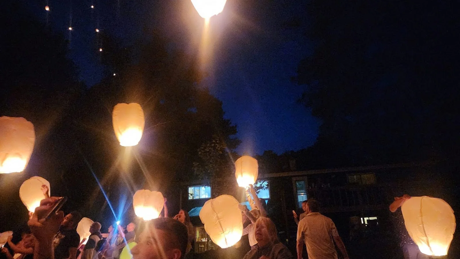 People releasing sky lanterns at night, illuminated by the lanterns' glow, against a dark blue sky and surrounding trees.