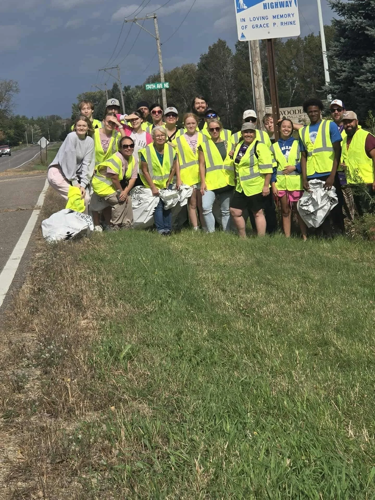 Group of people wearing bright yellow safety vests standing beside a road with a sign for Highway and 74th Ave NE in front of trees.