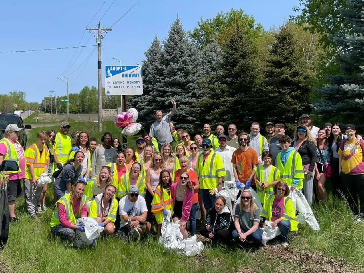 A large group of people participating in a community cleanup event, wearing safety vests and holding trash bags and balloons, gathered outdoors near a highway with trees in the background.