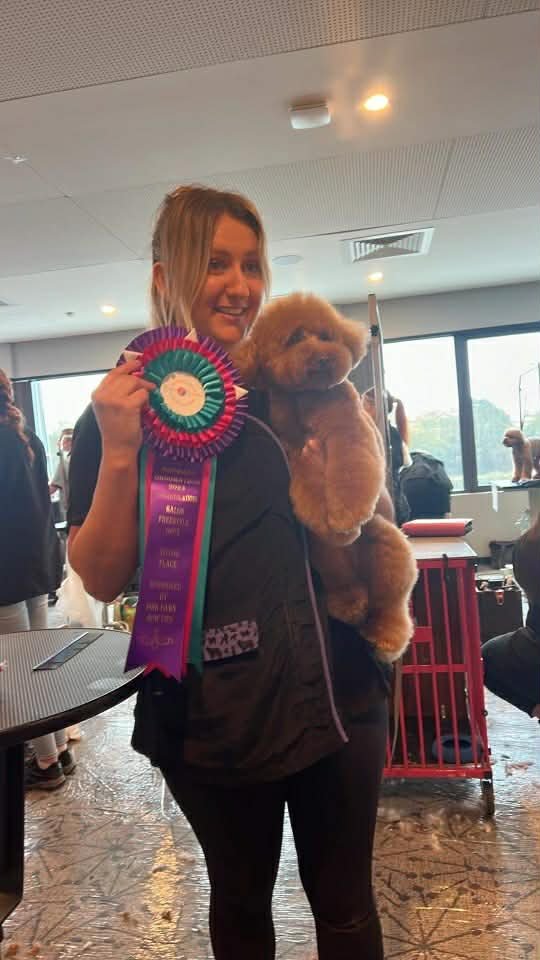 A woman smiling and holding a colorful award ribbon in one hand while carrying a small fluffy dog, possibly a poodle, in her other arm inside a well-lit room with large windows.