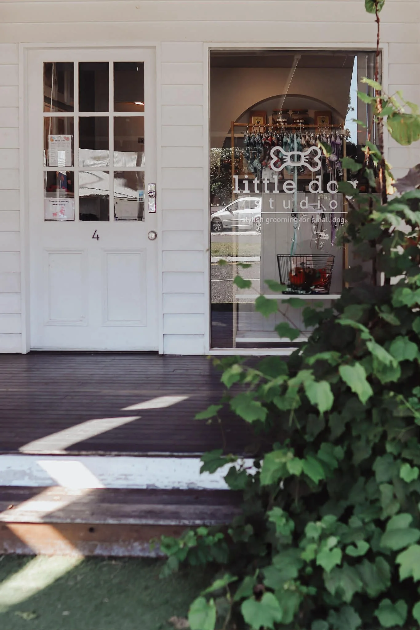 Front view of a small storefront with a white door labeled '4' on the left and a large window on the right displaying 'little do studios' with a dog bone logo above it. The window also indicates that it is a stylish grooming salon for small dogs. There is greenery in the foreground partially obscuring the view.