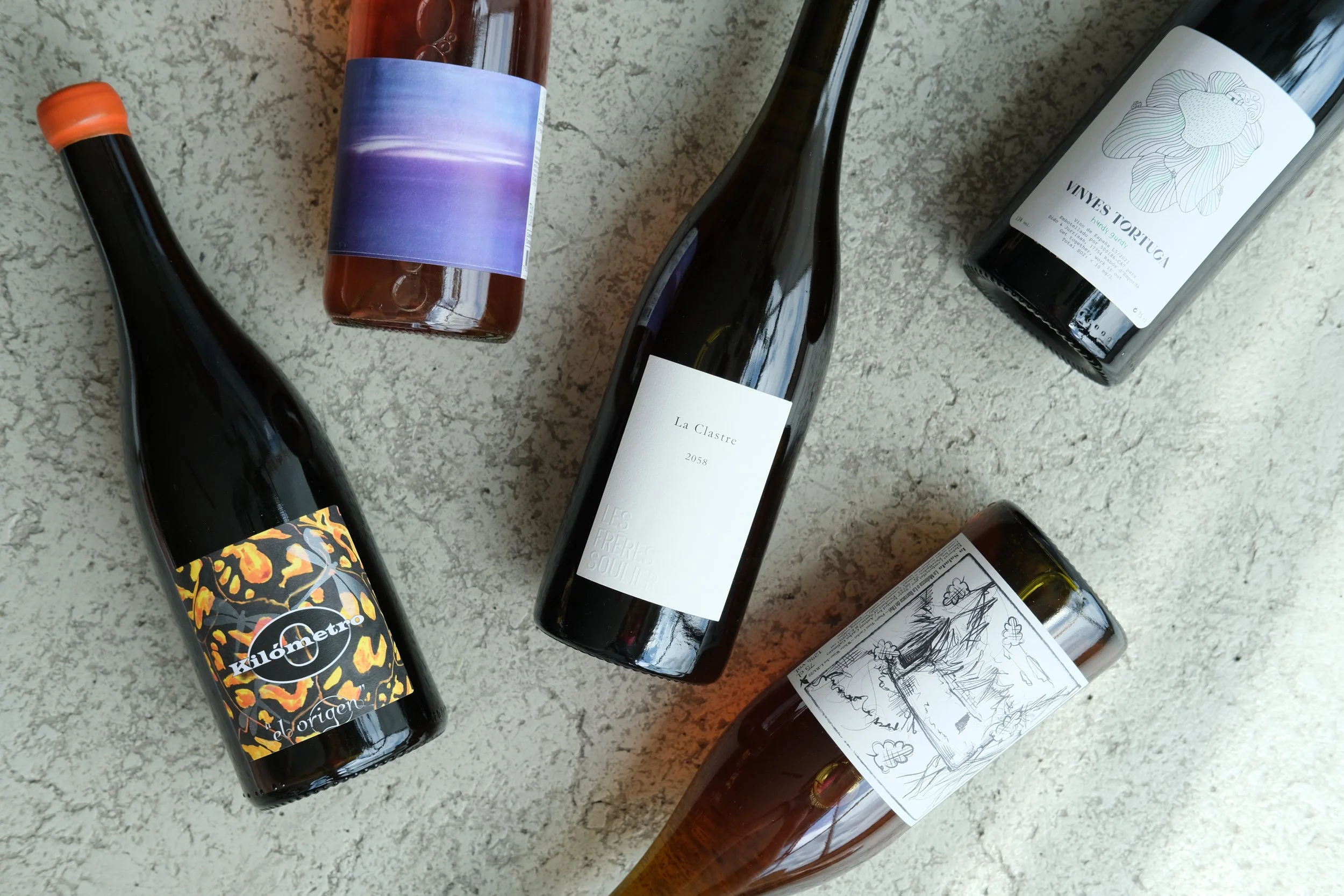 Five bottles of wine with various labels, laid out on a textured surface, including one with a plain white label, one with a black and white illustrated label, and others with colorful and minimalist designs.