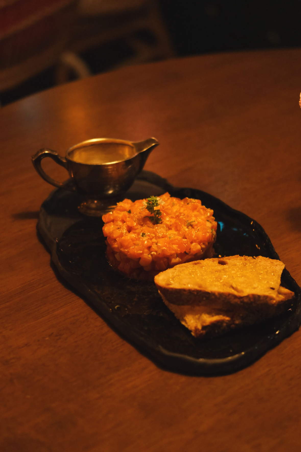 A black rectangular platter with a small serving of Indian-style chickpea curry, two pieces of toasted bread, and a small silver gravy boat on a wooden table.