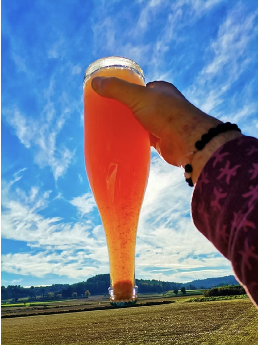 Person holding a tall glass of amber wine against a bright blue sky with scattered clouds, with a rural landscape below.