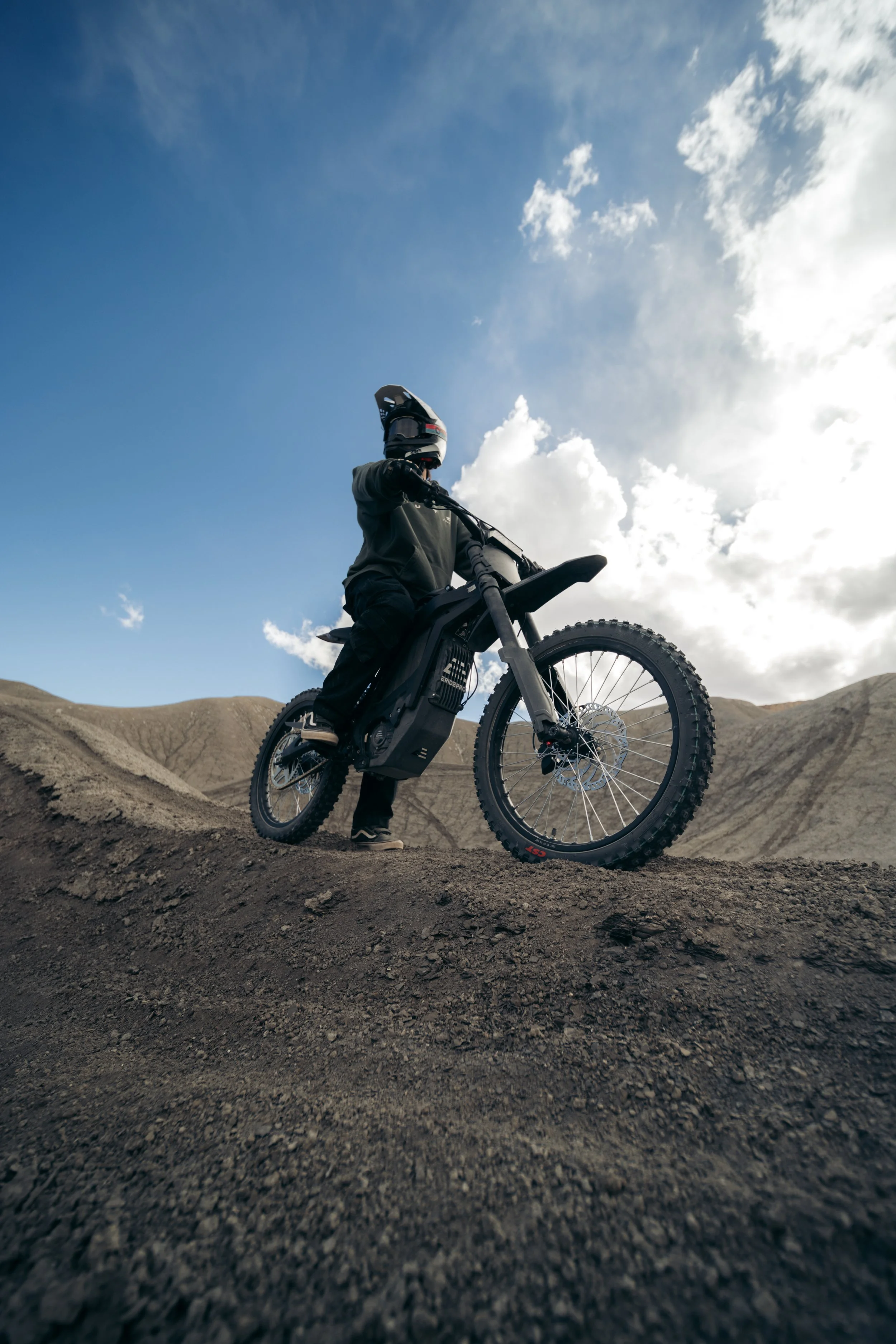 A person in protective gear and a helmet riding a black electric dirt bike on a dirt trail in a hilly landscape under a partly cloudy sky.